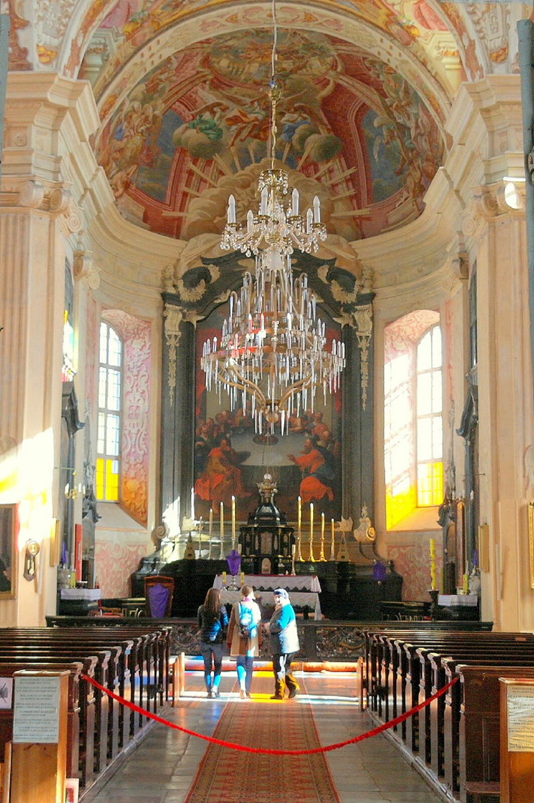 Ilustracja przedstawia wnętrze Kościoła Bożego Ciała w Nieświeżu. W kadrze znajduje się nawa główna, prezbiterium z ołtarzem i sklepienie apsydy pokryte freskami. W centrum fotografii znajduje się wielki, kryształowy żyrandol. We wnętrzu, po obu stronach, znajdują się drewniane ławki. Obszerna nawa i kopuła na skrzyżowaniu naw to także rozwiązania znane z Rzymu.