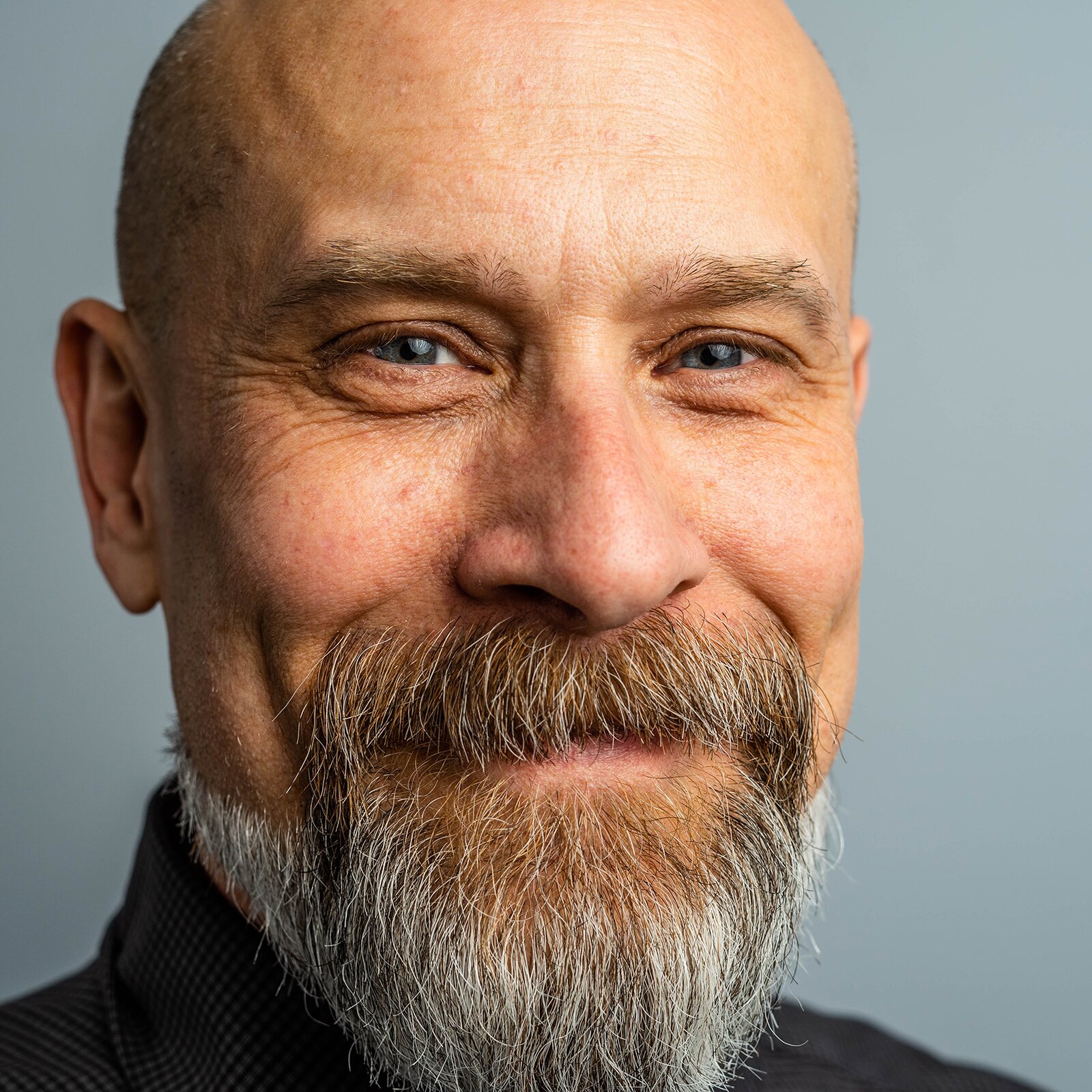 Zdjęcie przedstawia uśmiechniętego, starszego mężczyznę. Jest łysy. Ma niebieskie oczy i duży nos. Ma siwe wąsy i brodę.