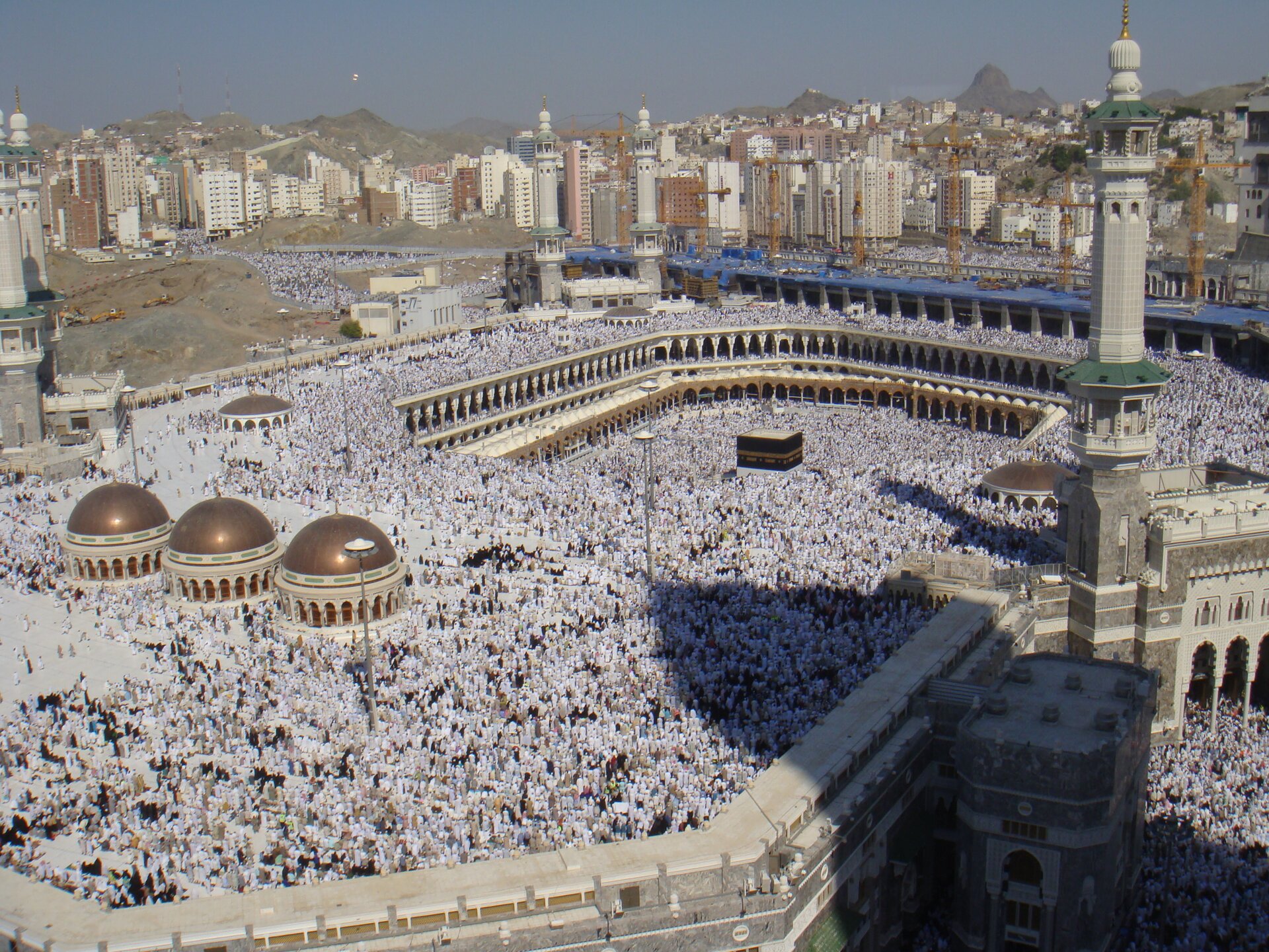 Zdjęcie przedstawia świątynię islamską z charakterystyczną wysoką wieżą, z której zwoływani są na modlitwę wierni. Przed nią oraz na jej dziedzińcu znajdują się tysiące modlących się ludzi.