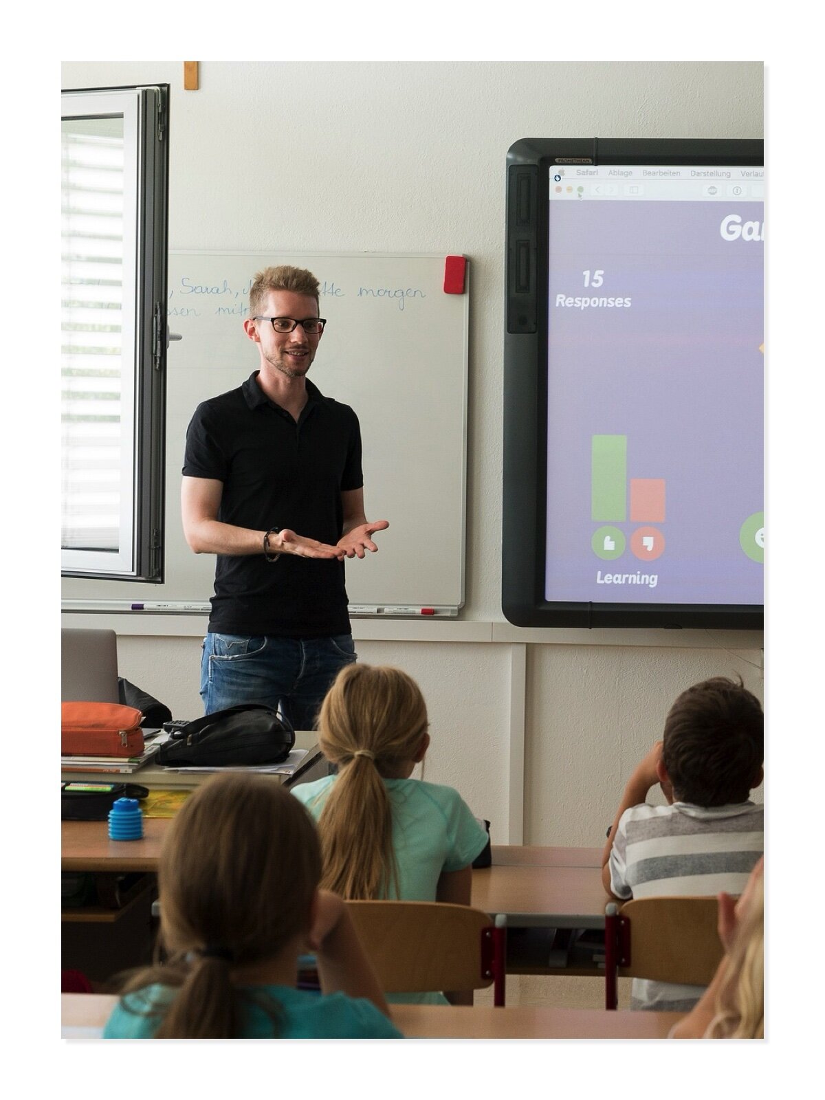 Fotografia przedstawia mężczyznę wykonującego zawód nauczyciela. Mężczyzna stoi przy tablicy interaktywnej. Przed nim w ławkach siedzą dzieci.
