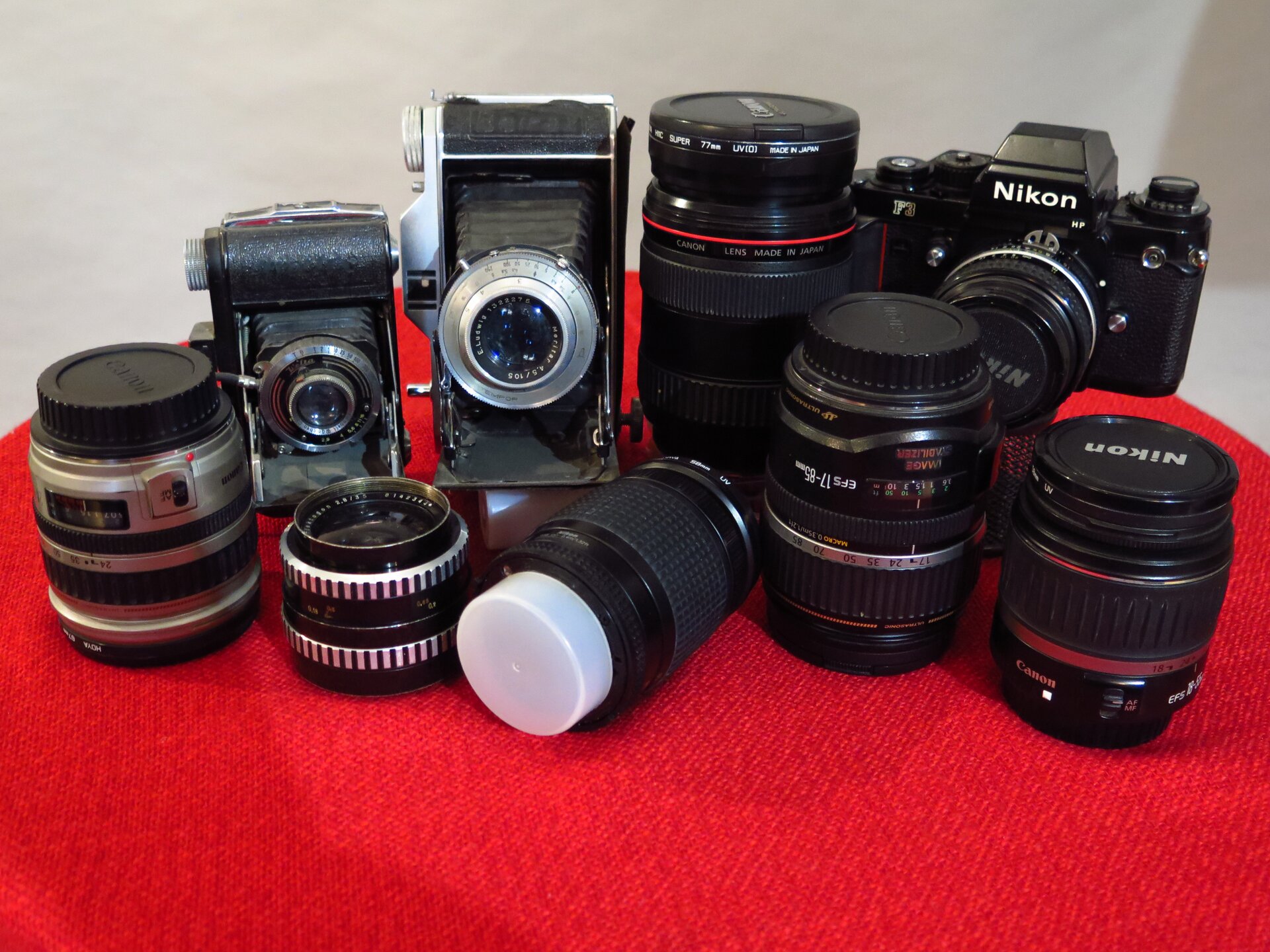 Zdjęcie przedstawia cyfrowe aparaty fotograficzne oraz różne modele obiektywów, ustawione obok sobie w artystycznym ułożeniu.