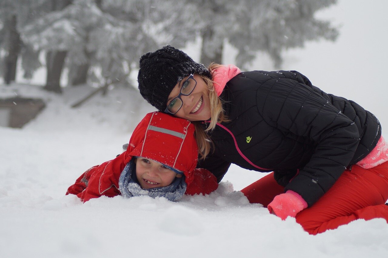 Mama z synem bawią się na śniegu. Chłopiec w czerwonej kurtce z kapturem na głowie leży na śniegu, obok mama klęczy i przytula go.