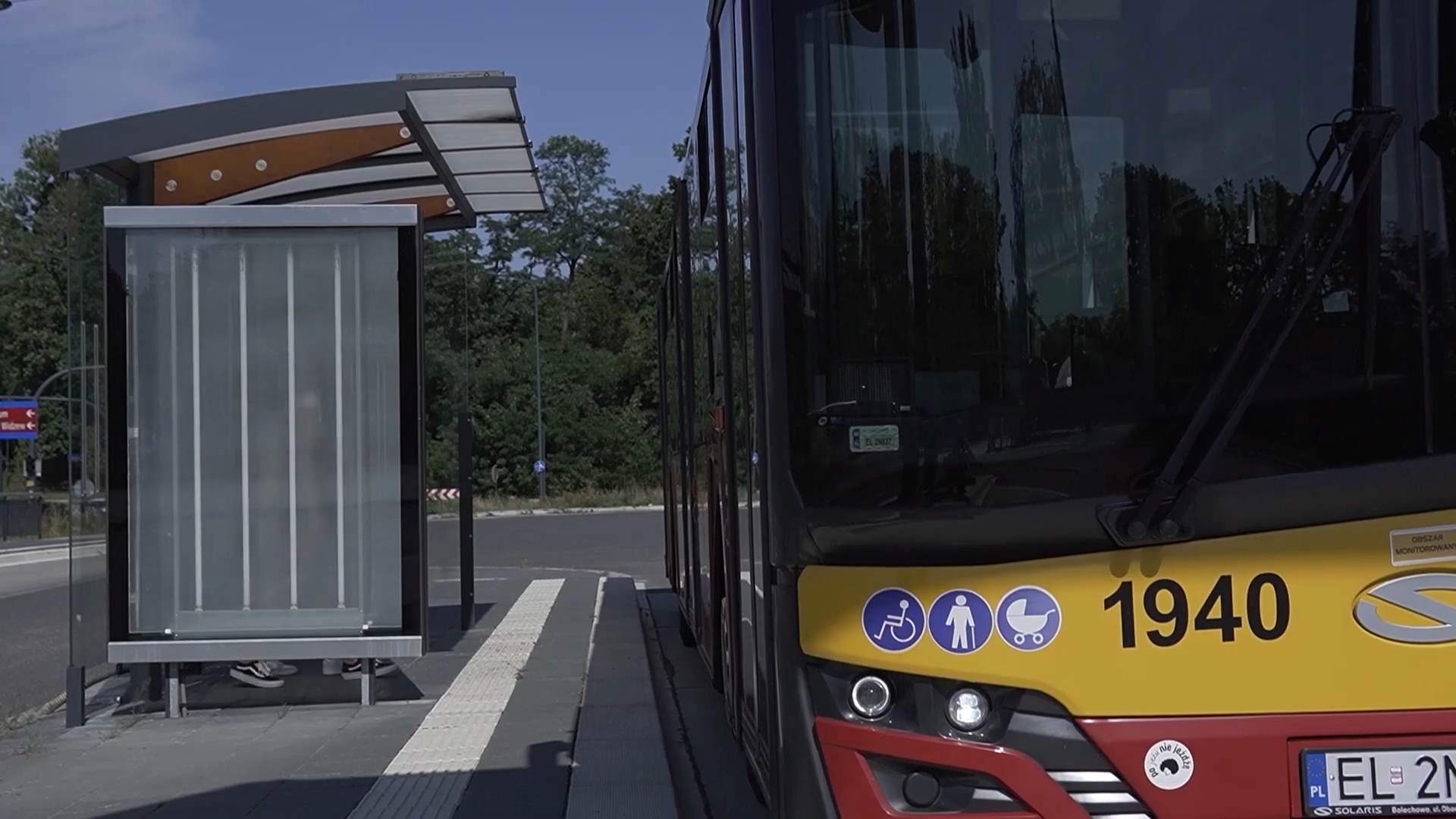 Zdjęcie przedstawia przystanek autobusowy, przy którym zatrzymał się autobus komunikacji miejskiej. W tle widoczna droga i drzewa.