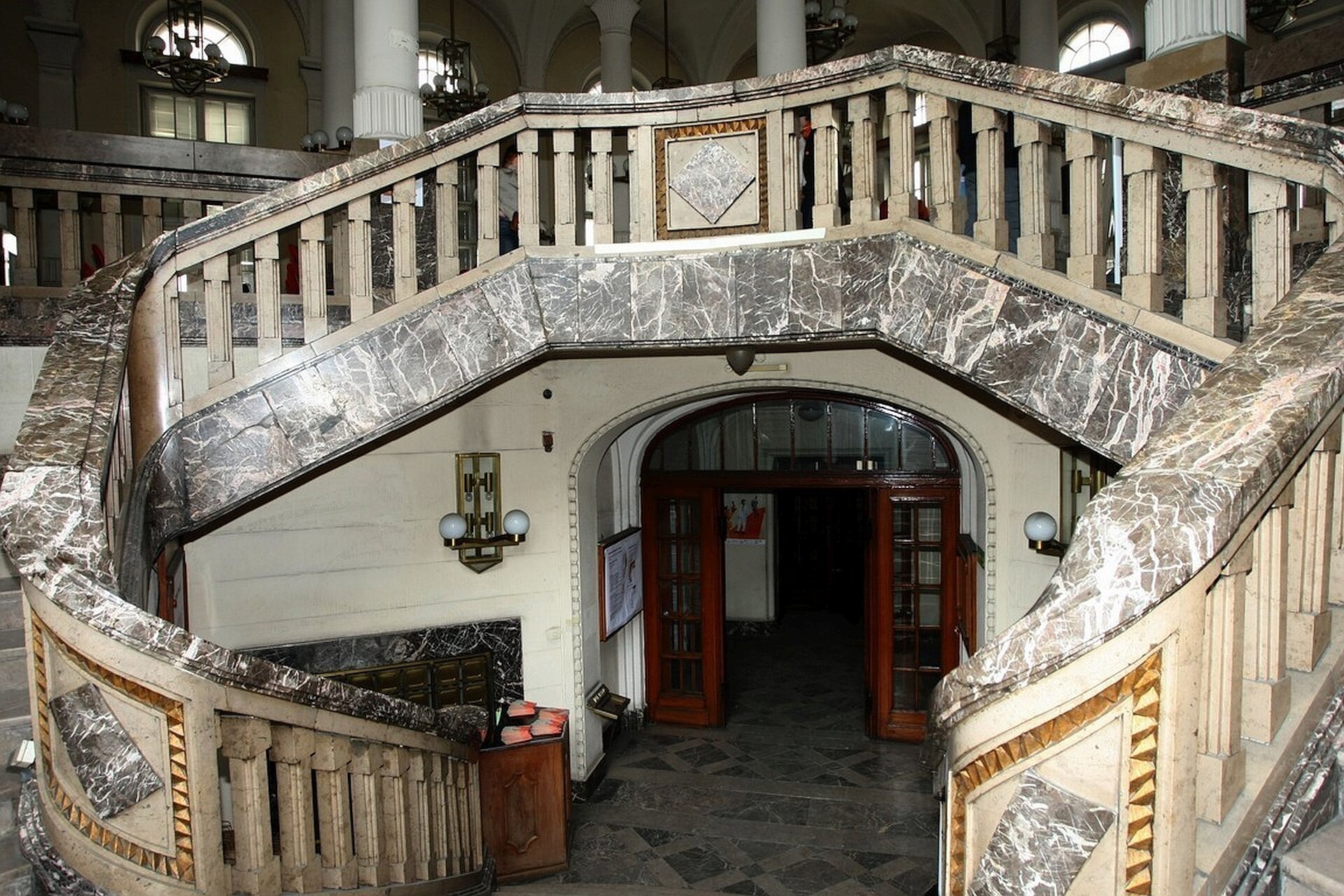 Ilustracja przedstawia Dawny Bank Polski, Gdynia, schody w stylu art déc. Widać przeszklone drzwi, które prowadzą do holu z szaro-czarną posadzką. z holu prowadzą zaokrąglone marmurowe schody. Wzdłuż schodów ciągną się marmurowe, gęsto ułożone obok siebie kolumny.