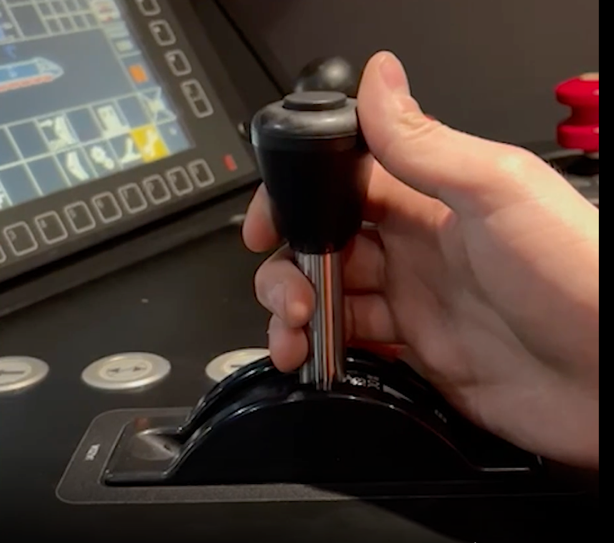 Zdjęcie przedstawia zadajnik jazdy i hamowania. Widoczna jest dźwignia z możliwością ruchu w przód i tył. Dłoń maszynisty trzyma dźwignię.