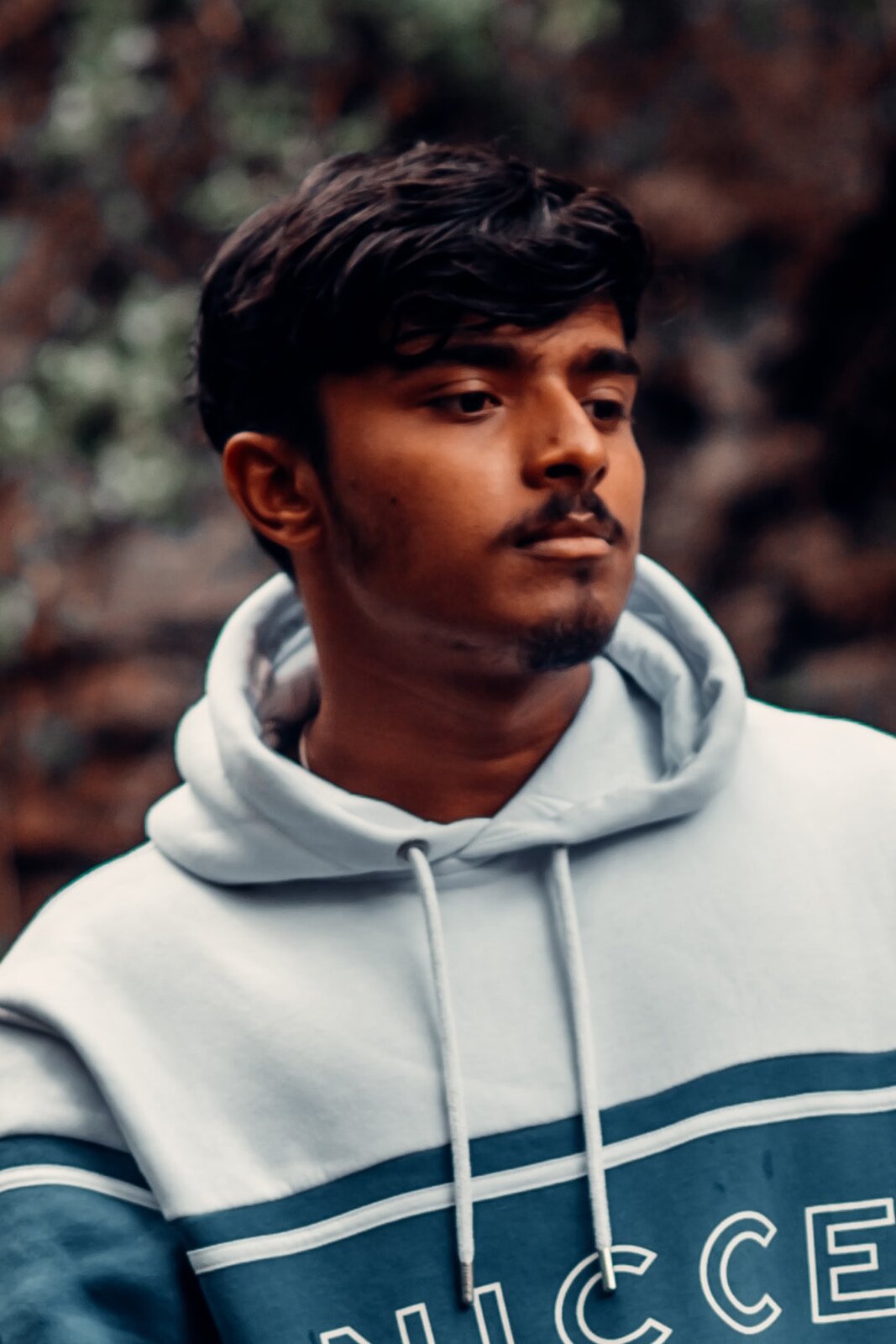 Zdjęcie przedstawia młodego Hindusa o śniadej karnacji, z czarnymi włosami i czarnym wąsem o ciemnych oczach. Ubrany jest w białą sportową bluzę z kapturem. 