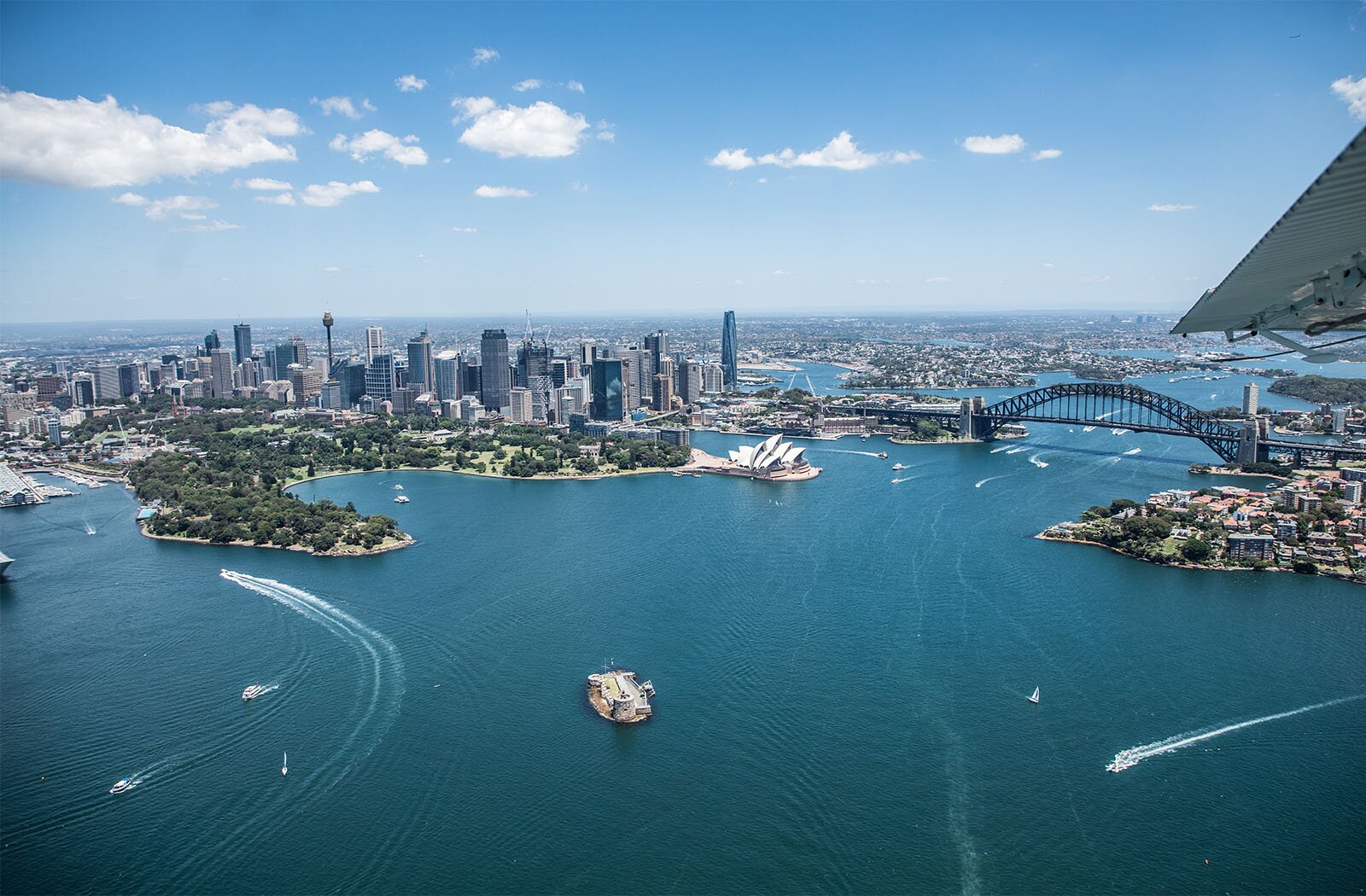 Zdjęcie przedstawia panoramę miasta Sydney w ujęciu z lotu ptaka. Na pierwszym planie widoczna jest tafla zbiornika wodnego z ciemnoniebieską wodą, licznymi zatokami i przystaniami. Na zbiorniku widoczne są płynące w różnych kierunkach statki, łodzie i motorówki. W głębi widoczny jest cypel porośnięty roślinnością. Po jego prawej stronie widoczny jest gmach opery, za którym widoczny jest most łączący dwa brzegi. W tle widoczne są wysokie wieżowce górujące nad miastem, a dalej rozciąga się miasto.