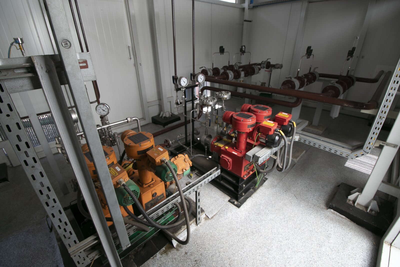 Na ekranie widoczne są pompy nurnikowe. Pompy nurnikowe stosowane są do wywarcia ciśnienia i wymuszenia przepływu płynów.Zdjęcie przedstawia cztery pompy nurnikowe. Dwie lewe są koloru pomarańczowego, a dwie prawe koloru czerwonego. Każda z pomp składa się z prostopadłościanu przy podstawie, z którego poprowadzony ku górze jest cylinder. U góry cylindra znajduje się walec o większym promieniu. Do cylindra dołączona jest pompa. Po przeciwnej stronie do walca dołączony jest kolejny walec, z którego poprowadzone są wąskie rurki. Od podstawy również poprowadzone są wąskie rurki. Rurki prowadzą do ciśnieniomierzy. Wszystkie pompy znajdują się na metalowym stelażu. W tle widoczne są rury.