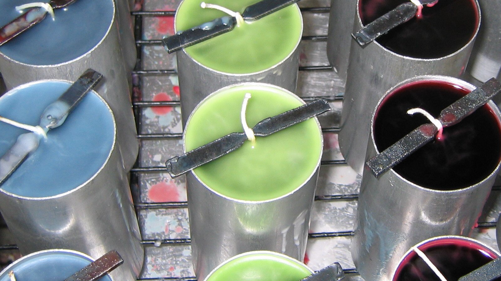 Zdjęcie przedstawia wysokie, cylindryczne, metalowe pojemniki, ułożone w trzech rzędach na metalowej kratce. Pojemniki wypełnione są woskiem w kolorze niebieskim, jasnozielonym i ciemnoczerwonym. Pośrodku każdego pojemnika z wosku wystaje biały knot przymocowany metalowym śmigiełkiem leżącym na krawędziach pojemnika.
