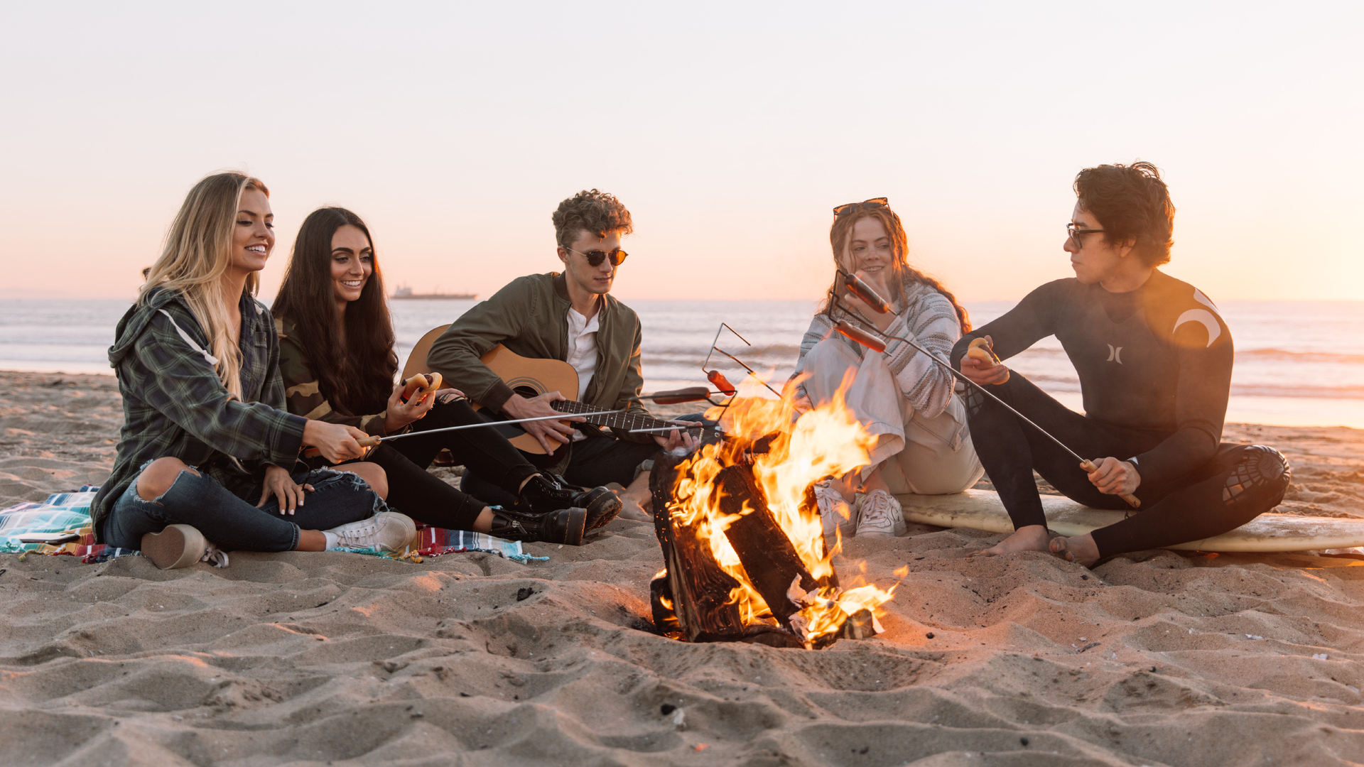 Zdjęcie przedstawia pięcioro młodych ludzi przy ognisku na plaży. Po lewej stronie przy ognisku siedzą dwie dziewczyny i trzymają kijki z kiełbasą nad ogniskiem. Pośrodku siedzi chłopak w ciemnych okularach z gitarą. Po prawej siedzą dziewczyna i chłopak. Trzymają również kijki z kiełbaskami nad ogniskiem.
