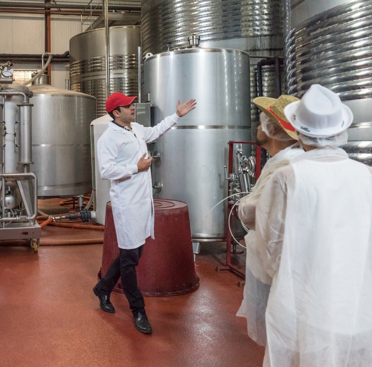 Zdjęcie prezentuje trzech mężczyzn we wnętrzu zakładu produkcyjnego. Jeden z mężczyzn wskazuje na stalowe silosy, które znajdują się w pomieszczeniu. Dwaj pozostali mężczyźni stoją i się przysłuchują.