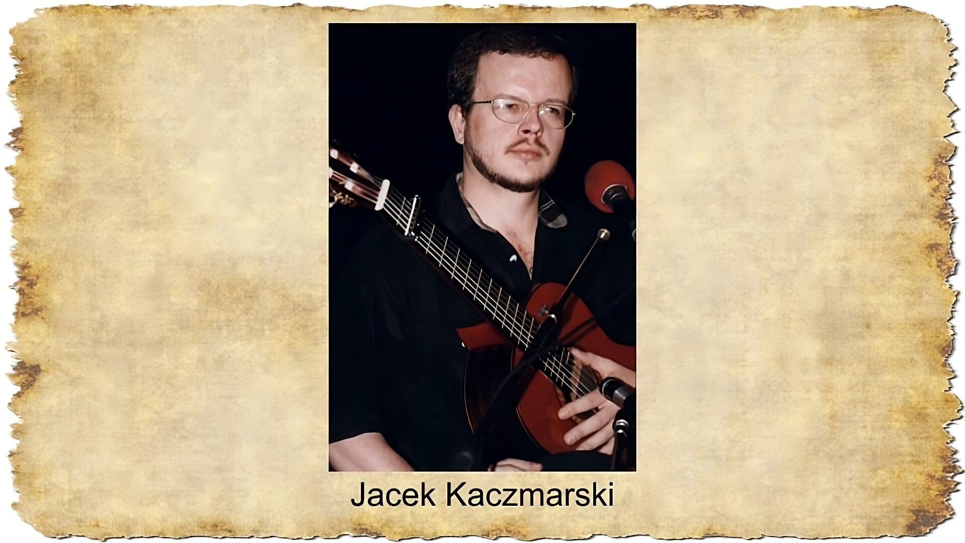 Po środku slajdu widoczny jest portret około czterdziestoletniego mężczyzny w okularach, o ciemnych włosach, brodzie i wąsach. Mężczyzna ubrany jest w czarną koszulę, w rękach trzyma gitarę, zaś przed nim stoi mikrofon. Tło fotografii jest czarne. Pod zdjęciem podpis: Jacek Kaczmarski.