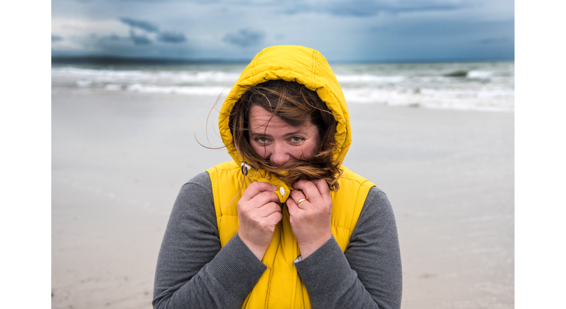 Zdjęcie przedstawia młodą kobietę na plaży nad morzem. Kobieta otula się kapturem kurtki. Morze jest wzburzone, na horyzoncie widoczne są burzowe chmury. 