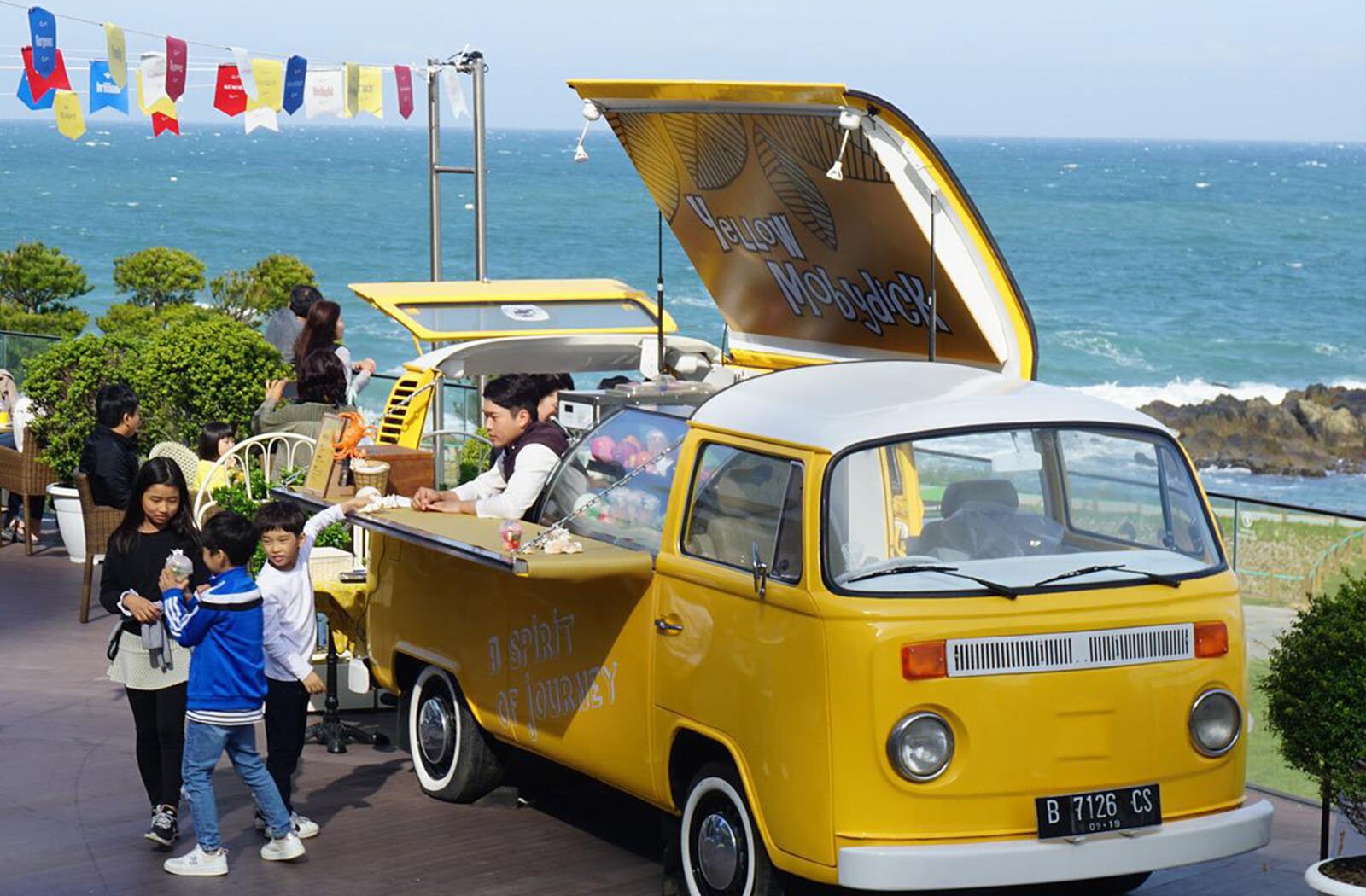 Zdjęcie przedstawia żółty food truck, który stoi na tarasie z widokiem na duży zbiornik wodny. Samochód ma otwarty dach i wysuniętą do przodu ladę. Na spodniej stronie dachu widoczny jest napis w języku angielskim: YELLOW MOBYDICK. W środku samochodu widoczny jest mężczyzna, który opiera się o blat lady i patrzy na trójkę dzieci stojących przed ladą. Dziewczynka spogląda na chłopca, których odchodzi od food trucka, a ręku trzyma jakiś pojemnik. Drugi chłopiec odwrócił do nich głowę, ale lewą ręką sięga po coś leżącego na ladzie. W tle, po lewej stronie, widoczne są osoby siedzące przy stolikach i stojące przy barierce tarasu, zwrócone w stronę wody.