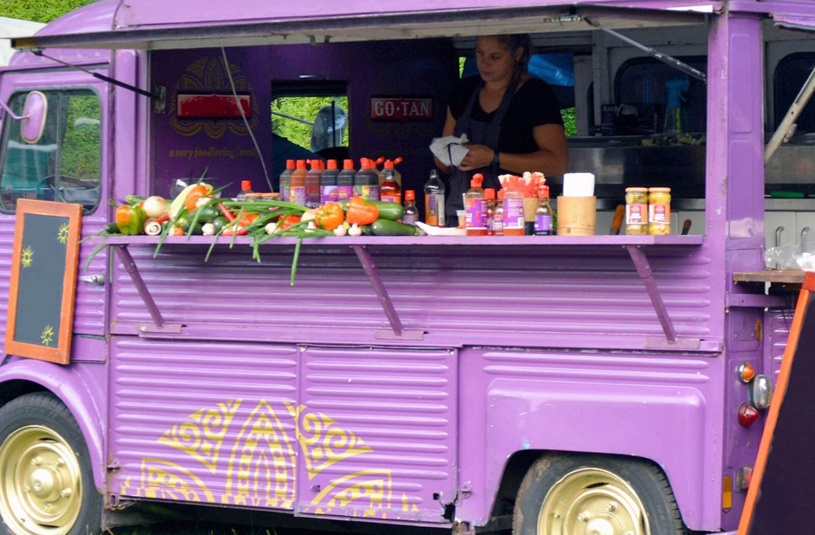 Zdjęcie przedstawia fioletowy samochód, przerobiony na obwoźny bar. Widoczna jest lada, na której stoją butelki z sokami i sosami, słoiki z piklami, pojemniki z serwetkami i sztućcami oraz leżą różne warzywa (pomidory, ogórki, czerwona papryka, cukinia, szczypior). Wewnątrz, za ladą widoczna jest stojąca kobieta, która przeciera białą ściereczką okulary. Na prawo i lewo za nią widoczne jest wyposażenie kuchni.