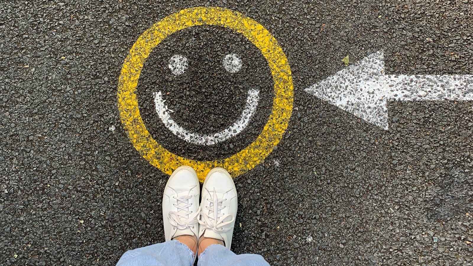 Zdjęcie przedstawia namalowany na asfalcie znak uśmiechniętej buźki z głową w kolorze żółtym oraz oczami i ustami w kolorze białym. Z prawej strony widoczna jest biała strzałka skierowana w stronę buźki. W dolnej części znaku widoczne stopy w białych sportowych butach. 