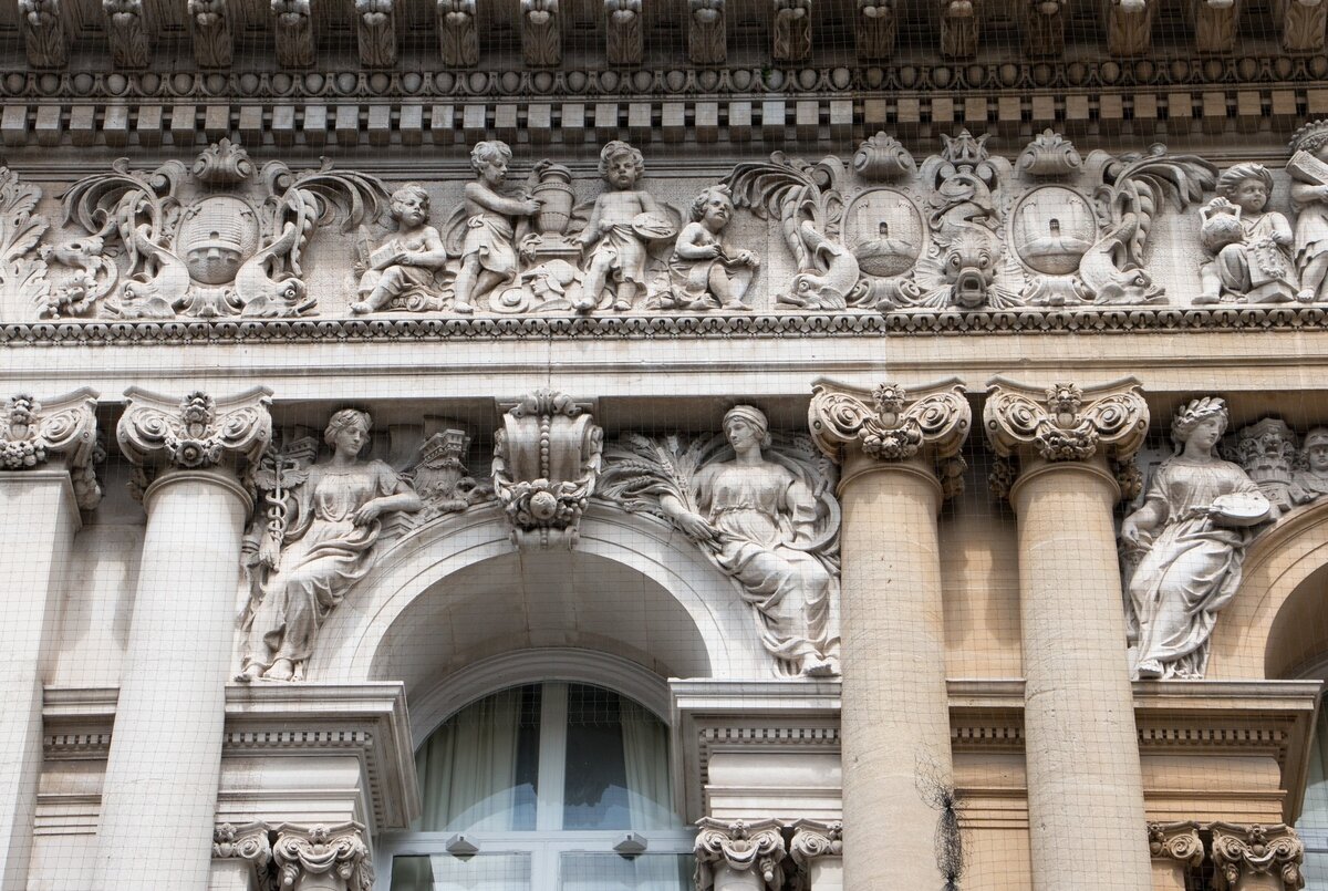 Zdjęcie przedstawia bogato zdobioną rzeźbami fasadę budynku. Okna są zakończone kamiennymi łukami. Nad oknami umieszczono rzeźby kobiet w długich szatach. Jedna z nich trzyma laskę eskulapa. Okna są poprzedzielane gładkimi kolumnami. Nad kolumnami znajduje się wystający poza lico ściany gzyms, a nad nim kolejne rzeźby, przedstawiające dzieci oraz dekoracje roślinne.