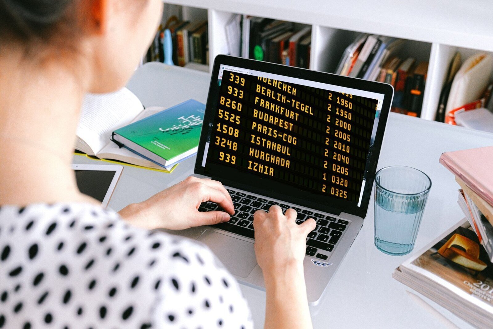 Zdjęcie przedstawia fragment postaci siedzącej przy biurku, na którym widoczny jest otwarty laptop. Osoba trzyma ręce na klawiaturze laptopa. Na monitorze laptopa widoczne są propozycje lotów ułożone w trzech kolumnach: numer, nazwa miasta i numery. Widoczne są nazwy: BERLIN‑TEGEL, FRANKFURT, BUDAPEST, PARIS‑CDG, ISTANBUL, HURGHADA, IZMIR. Po prawej stronie laptopa na biurku stoi szklanka z wodą i stos książęk, zaś po lewej stronie otwarty notes, książka i tablet. W tle widoczny jest regał z książkami.