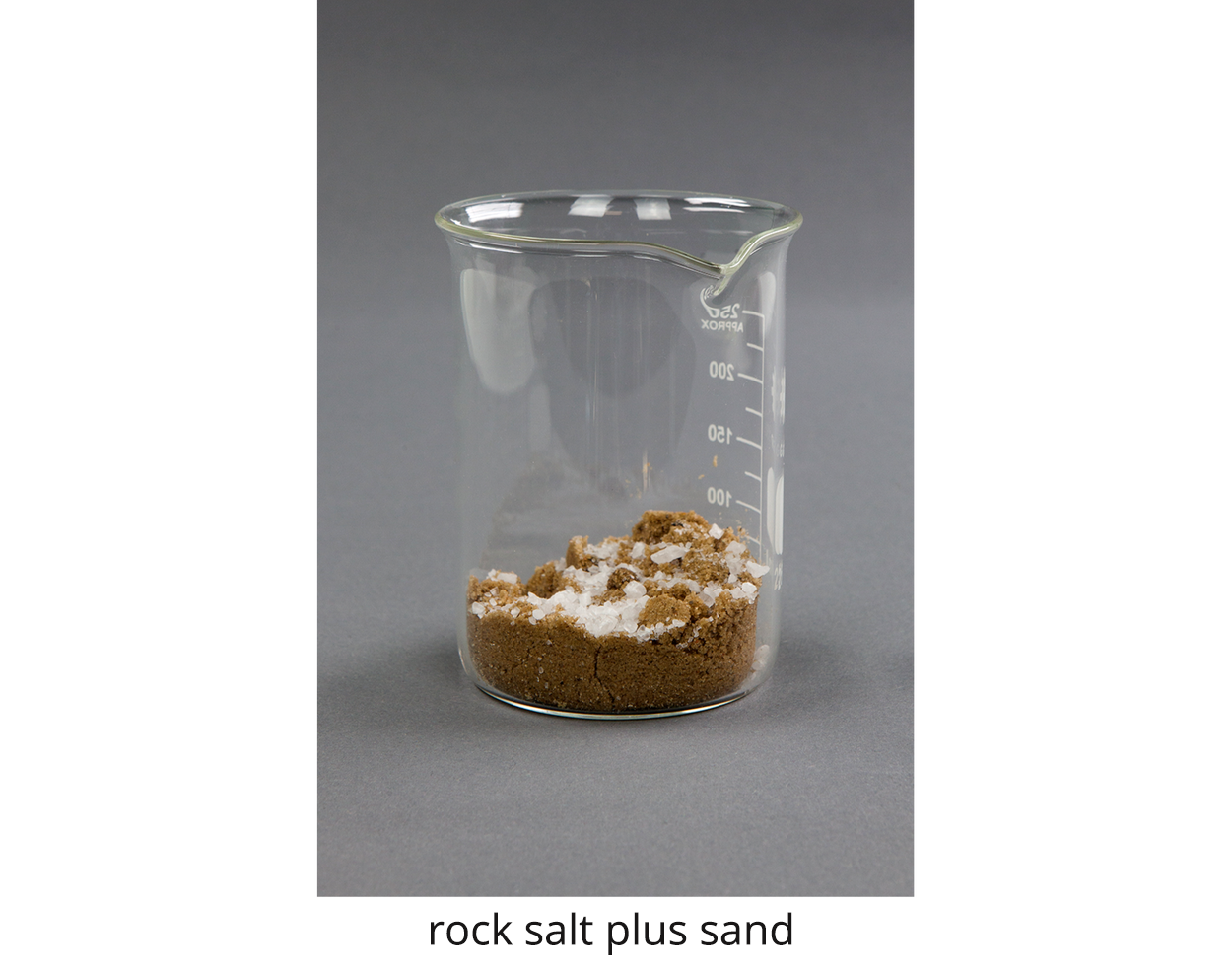 Zdjęcie przedstawia piasek i kryształ białej soli, rock salt plus sand w zlewce. Sól jest rozsypana na beżowym piasku. Substancje nie są zmieszane.