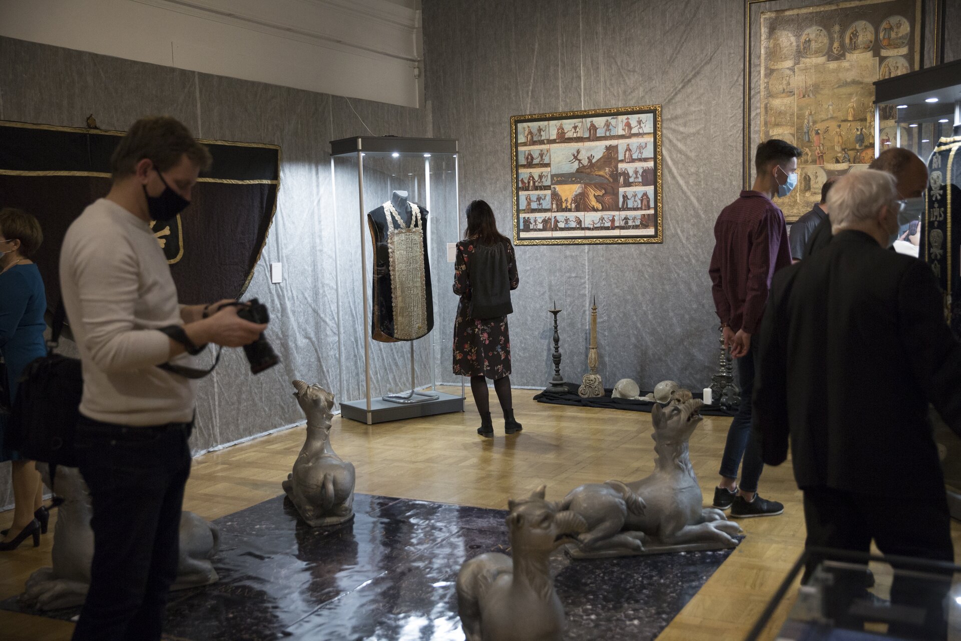 Zdjęcie ukazuje wnętrze galerii lub sali muzealnej gdzie prezentowana jest kolekcja dzieł sztuki. Na środku pomieszczenia stoją rzeźby zwierząt. W pomieszczeniu stoi wiele osób podziwiających eksponaty. Na środku fotografii ukazana jest ściana, na której wisi obraz, tuż obok niego po lewej stronie stoi gablota, za którą wisi czarna szata zdobiona beżowymi i srebrnymi ozdobami. Przed gablotą stoi kobieta. 