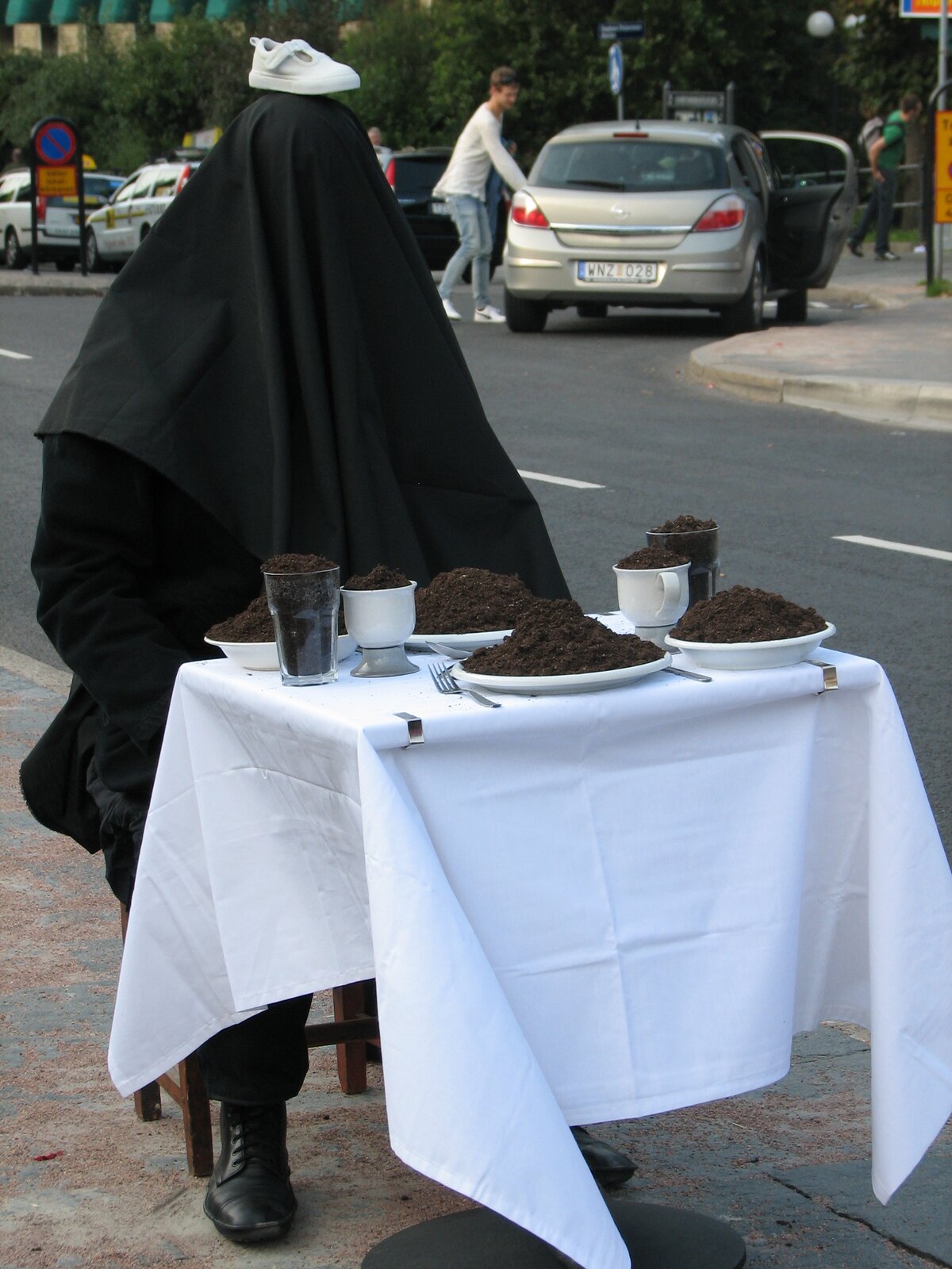 Zdjęcie przedstawia dzieło Alastaira MacLennana [czyt. alistera meklenena] „Performance [czyt. performans] artystyczny”. Na chodniku, przy ulicy stoi stolik przykryty białym obrusem. Na stoliku stoją cztery talerze, dwie filiżanki i dwie szklanki. Naczynia wypełnione są czarną ziemią. Przy stoliku, na krześle siedzi mężczyzna ubrany na czarno. Na głowę ma założony czarny materiał, który całkowicie zasłania jego twarz i ramiona. Dodatkowo na czubku głowy leży biały sportowy but.