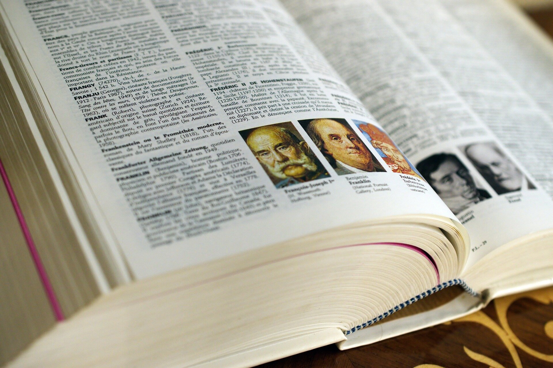 Zdjęcie przedstawia otwartą encyklopedię. Widoczne są hasła wraz z definicjami oraz niewielkie ilustracje przedstawiające portrety mężczyzn. Lewa strona jest czytelna i ostra, zaś prawa jest lekko zamglona.