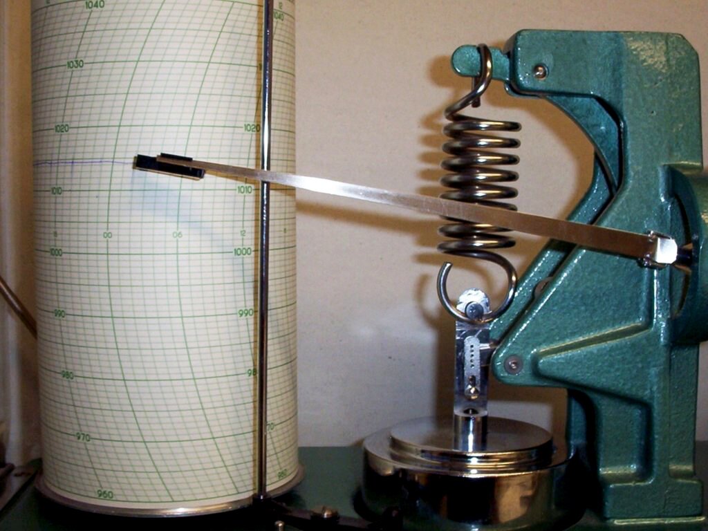 Fotografia przedstawiająca barograf. Jest to plastikowe, przezroczyste naczynie z pokrywką, na której jest metalowa rączka do przenoszenia. W środku naczynia widać: po lewej stronie rolkę papieru stojącą pionowo, po prawej metalowy mechanizm z trzema wystającymi prętami, które są ruchome, a ich zakończenia dotykają rolki papieru po lewej stronie. 