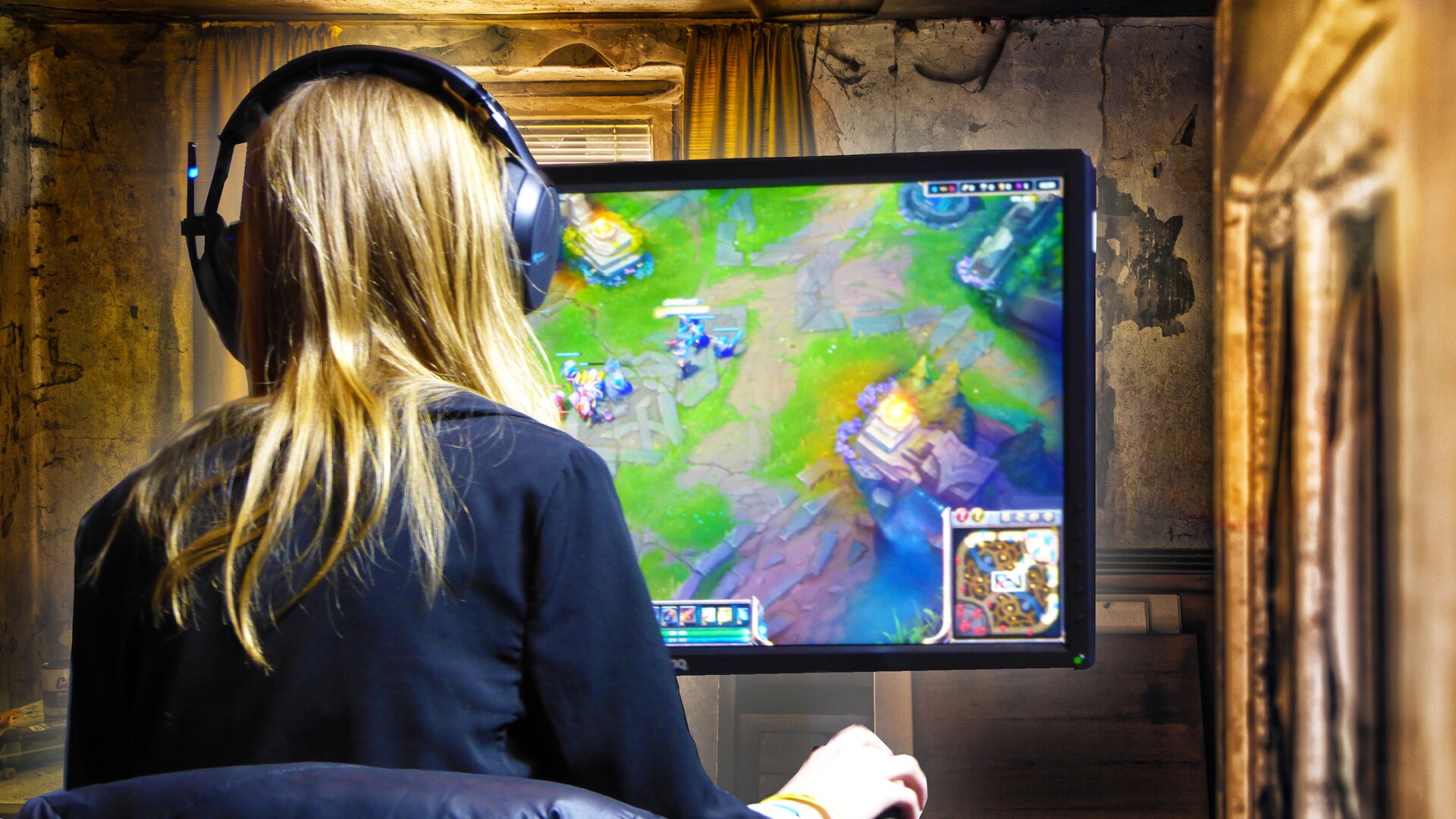 Ilustracja przedstawia dziewczynę siedzącą przed komputerem, grającą w grę komputerową. Dziewczyna ma długie blond włosy i słuchawki na uszach. Przed nią stoi duży monitor, gdzie wyświetlana jest gra, prawdopodobnie strategiczna. Dziewczyna przebywa w pomieszczeniu, które sugeruje, że jest częścią tej gry.