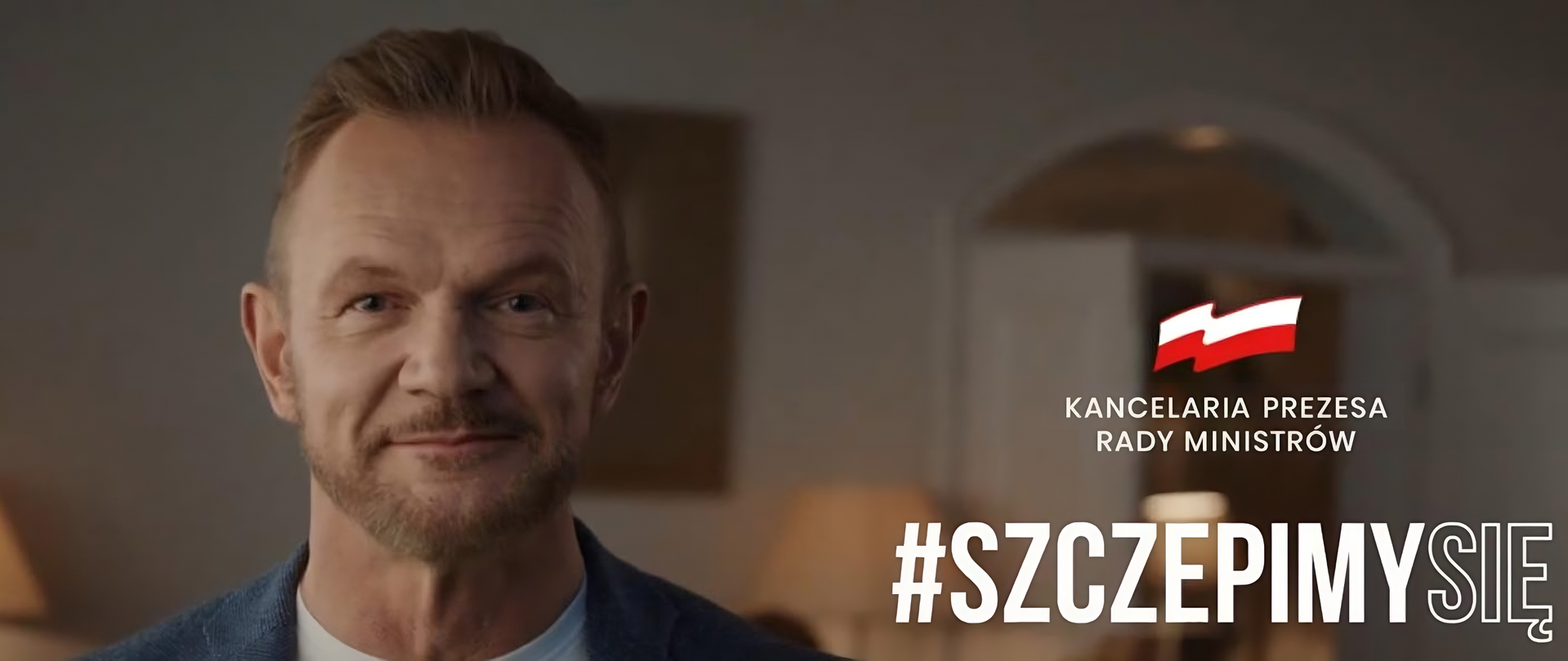 Kliknij żeby powiększyć Na zdjęciu po lewej stronie jest głowa aktora Cezarego Pazury, który delikatnie się uśmiecha. Po lewej stronie jest niewielka flaga Polski. Pod nią znajduje się logotyp Kancelarii Prezesa Rady Ministrów z flagą Polski i napisem Kancelaria Prezesa Rady Ministrów. W prawym dolnym rogu jest duży napis: #szczepimy się.
