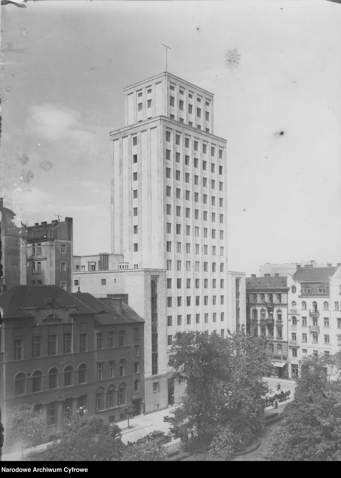 Zdjęcie przedstawia jasny, wysoki wieżowiec. Wokół niego znajduje się o wiele niższa miejska zabudowa.