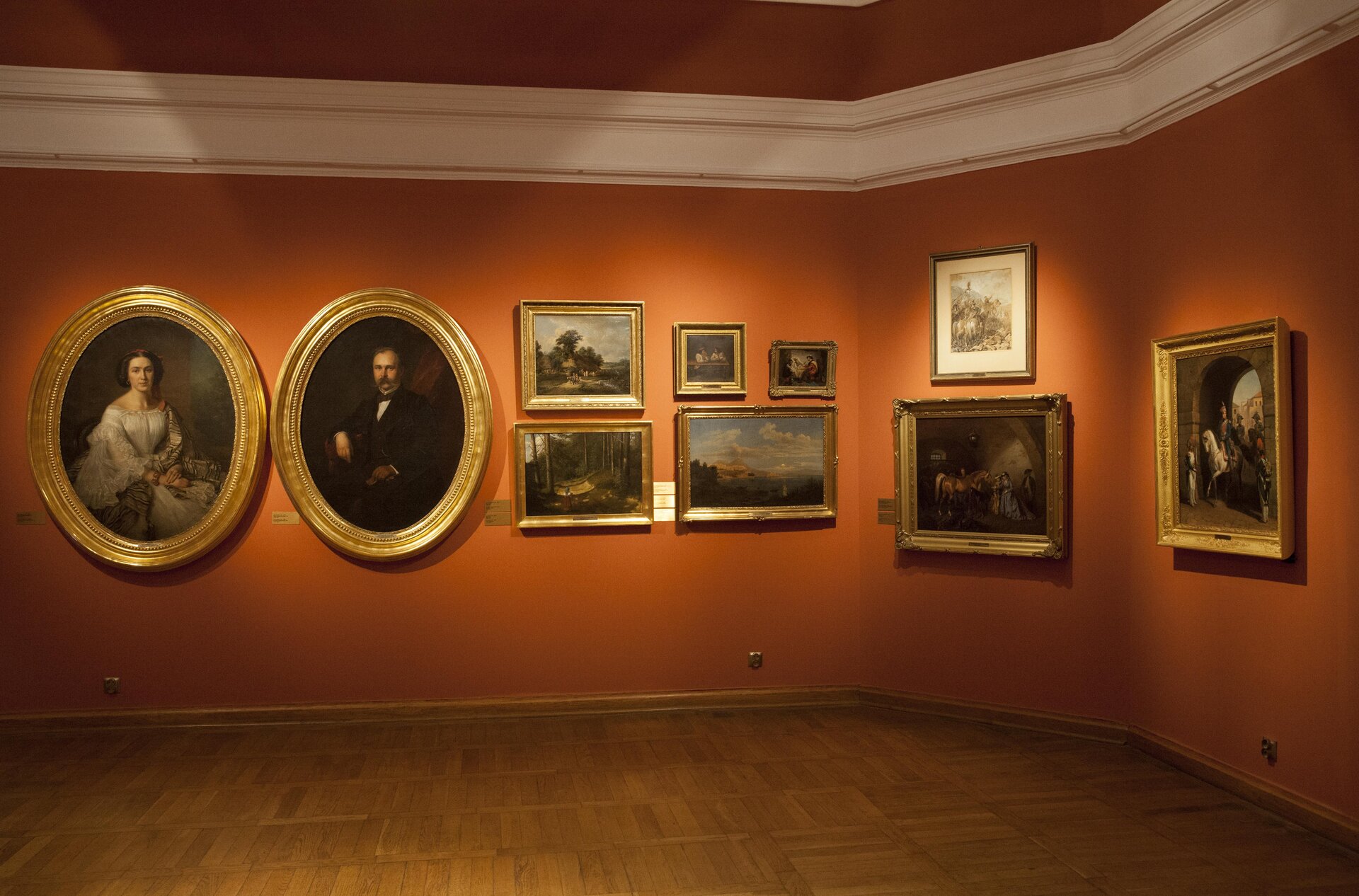 Zdjęcie przedstawia fragment pokoju z wystawą obrazów. Na tle pomarańczowej ściany wiszą obrazy w złotych ramach. Po lewej stronie znajdują się dwa owalne obrazy, na jednym jest kobieta, a na drugim mężczyzna. Dalej wiszą prostokątne obrazy, przedstawiające pejzaże. Po prawej stronie wiszą trzy obrazy. Na jednym jest mężczyzna na białym koniu, przejeżdżający przez bramę. Inny przedstawia konia i kilka osób w ciemnym pomieszczeniu.