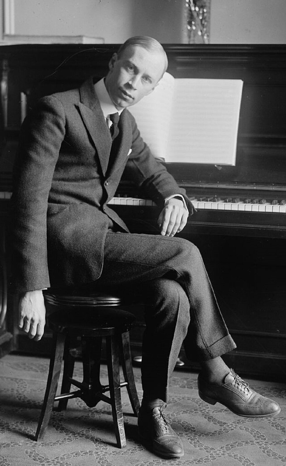 Ilustracja przedstawia Sergiusza Prokofiewa, który był rosyjskim kompozytorem, pianistą-wirtuozem oraz dyrygentem, jednym z najważniejszych twórców awangardy pierwszej połowy XX w. Mężczyzna w średnim wieku siedzi przy pianinie. Ubrany elegancko w szary garnitur i białą marynarkę.