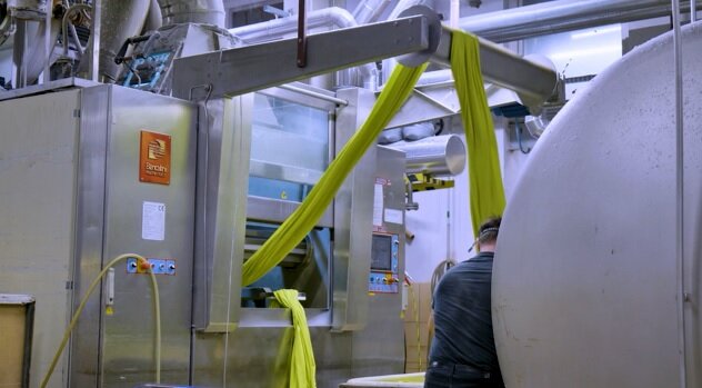 Zdjęcie przedstawia bielnik. W obudowie maszyny jest otwarte okno. Wewnątrz widoczny jest wał oraz żółta tkanina. Po lewej stronie obudowy są przyciski sterujące. Po prawej stronie obudowy jest ekran panelu sterującego oraz przyciski. Urządzenie podłączone jest do rur o różnej średnicy. Przy walcowatym pojemniku stoi pracownik. 