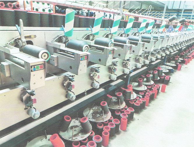 Zdjęcie przedstawia przewijarkę. Maszyna składa się z licznych, takich samych stanowisk przewijania. Na każdym stanowisku zamontowana jest szpulka szarych nici. Na maszynie i pod maszyną stoją inne szpulki. 