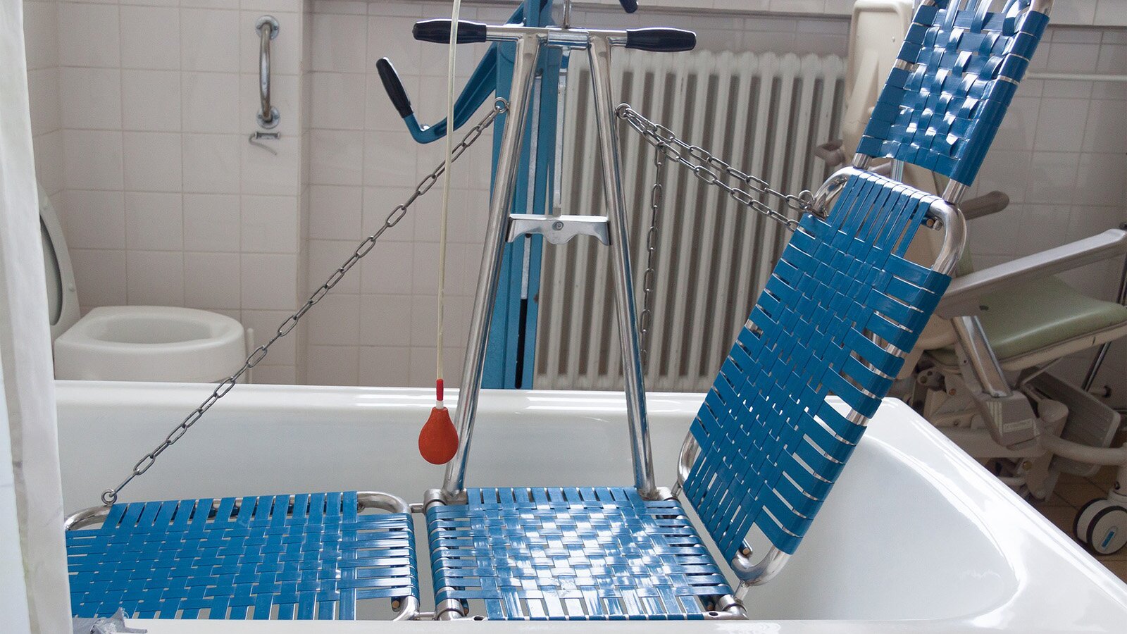Zdjęcie przedstawia leżak kąpielowy dla osób z niepełnosprawnościami, który – za pomocą łańcuchów – jest podwieszony na metalowym stelażu w wannie. Leżak wykonany jest z plecionki w kolorze niebieskim, jego poszczególne części są składane, a zaczep przy oparciu sugeruje możliwość regulacji tego elementu.