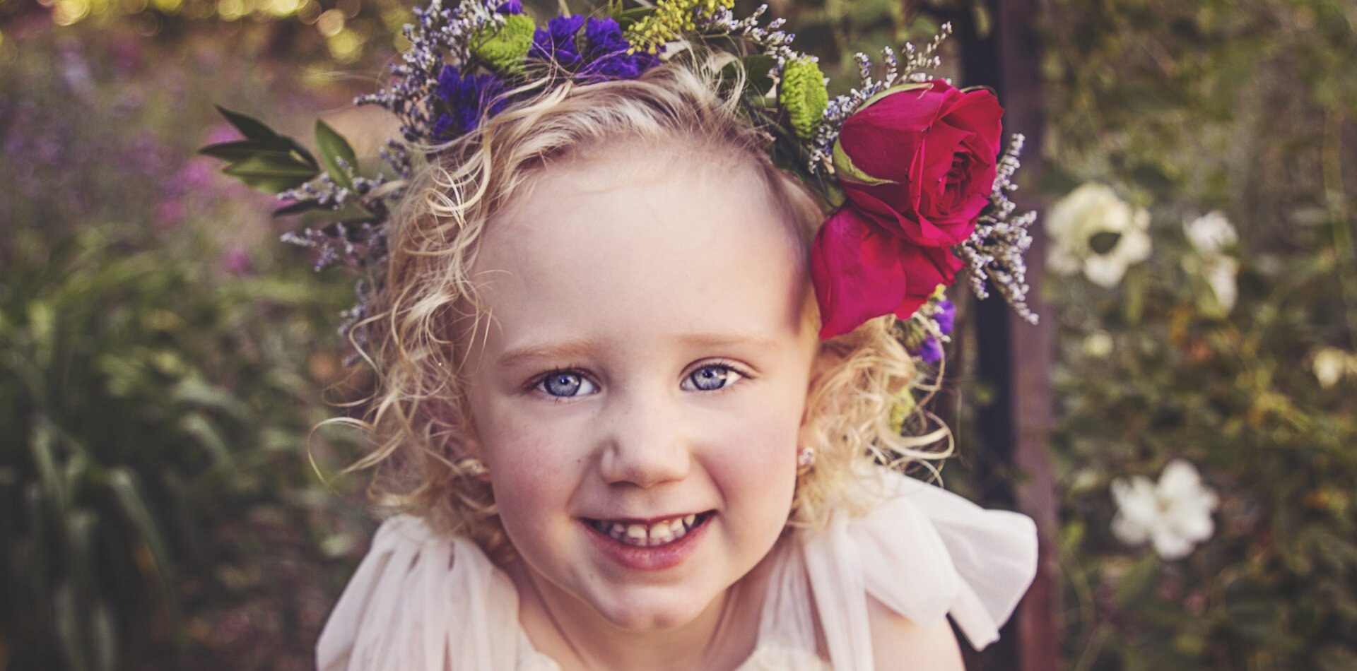 Fotograficzny portret uśmiechniętej dziewczynki o jasnych, lokowanych włosach. Na głowie widać wianek z dopiętymi dwiema dużymi, czerwonymi różami. W tle jest nieostro ukazany ogród kwiatowy.