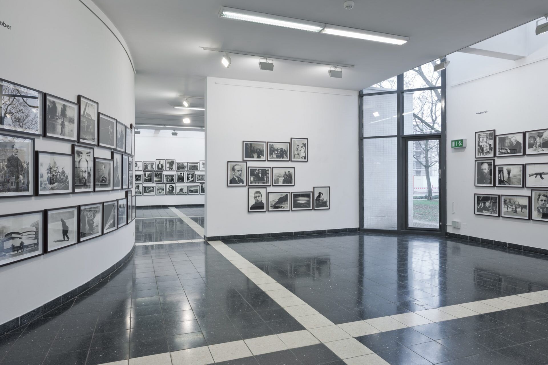 Zdjęcie przedstawia nowoczesny hol w budynku, gdzie na wszystkich ścianach zaaranżowano wystawę fotografii. Wszystkie fotografie są bez koloru, mają podobne wielkości, są oprawione w ramki. Zostały zawieszone na jasnych ścianach w kilku rzędach.  