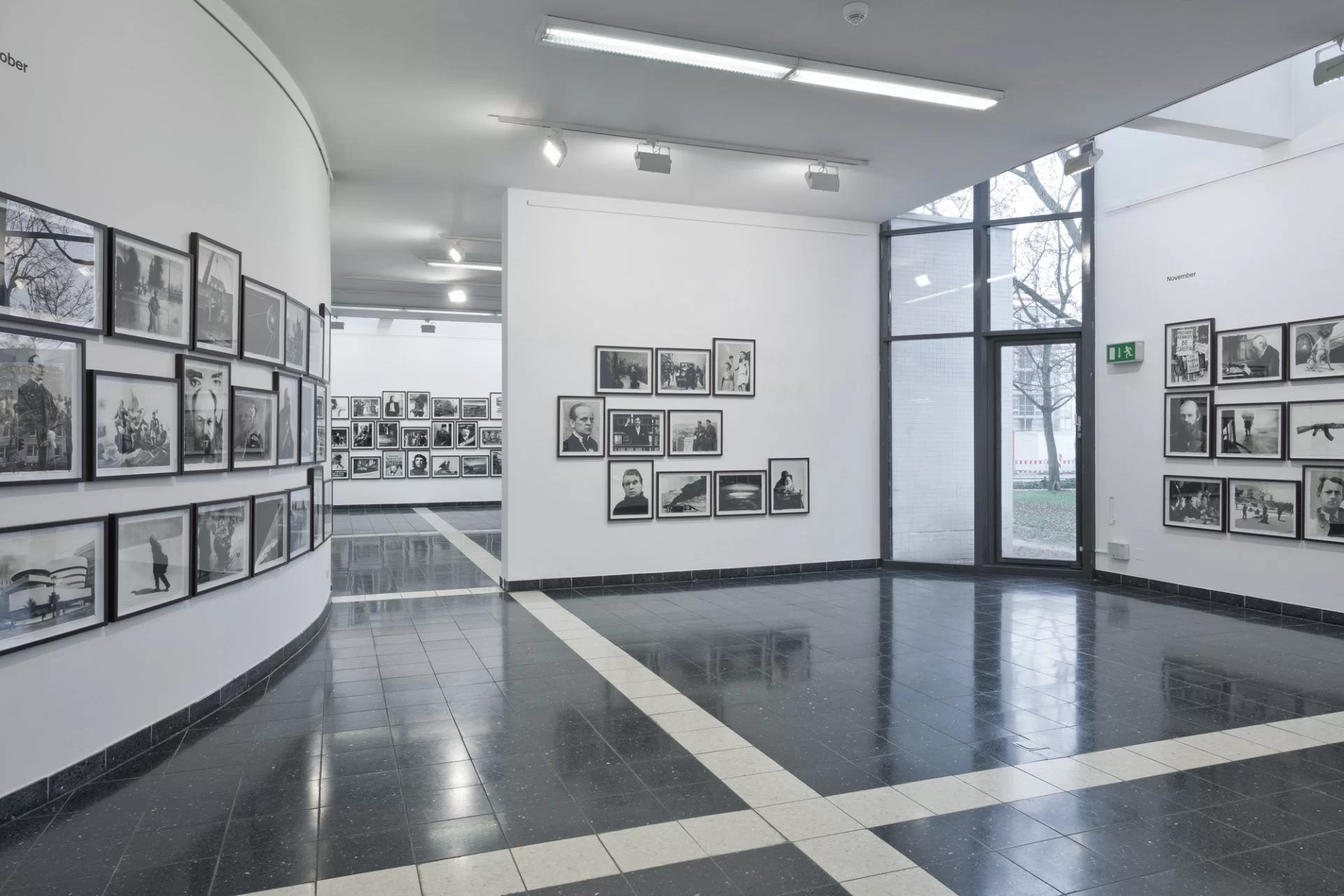Zdjęcie przedstawia nowoczesny hol w budynku, gdzie na wszystkich ścianach zaaranżowano wystawę fotografii. Wszystkie fotografie są bez koloru, mają podobne wielkości, są oprawione w ramki. Zostały zawieszone na jasnych ścianach w kilku rzędach.  