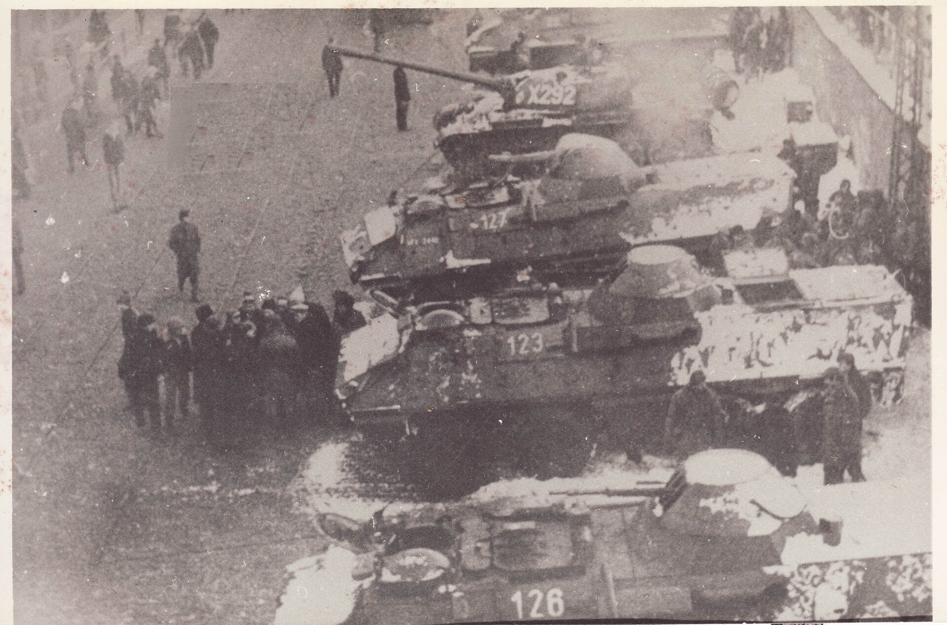 Czarno‑białe zdjęcie przedstawia widok na ulicę. Po prawej widać cztery stojące obok siebie czołgi, częściowo obsypane śniegiem. Po lewej stronie obok czołgów widać skupionych w grupach ludzi.
