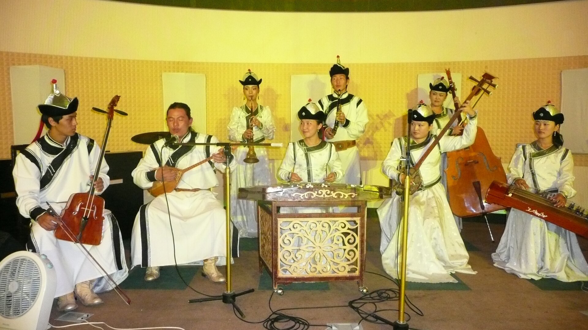 Na zdjęciu grupa muzyków. Mają na sobie białe stroje oraz czarne kapelusze. Na środku znajduje się kobieta grająca na instrumencie perkusyjnym. Wokół osoby grające na instrumentach strunowych. W tyle dwie osoby grające na instrumentach dętych. 