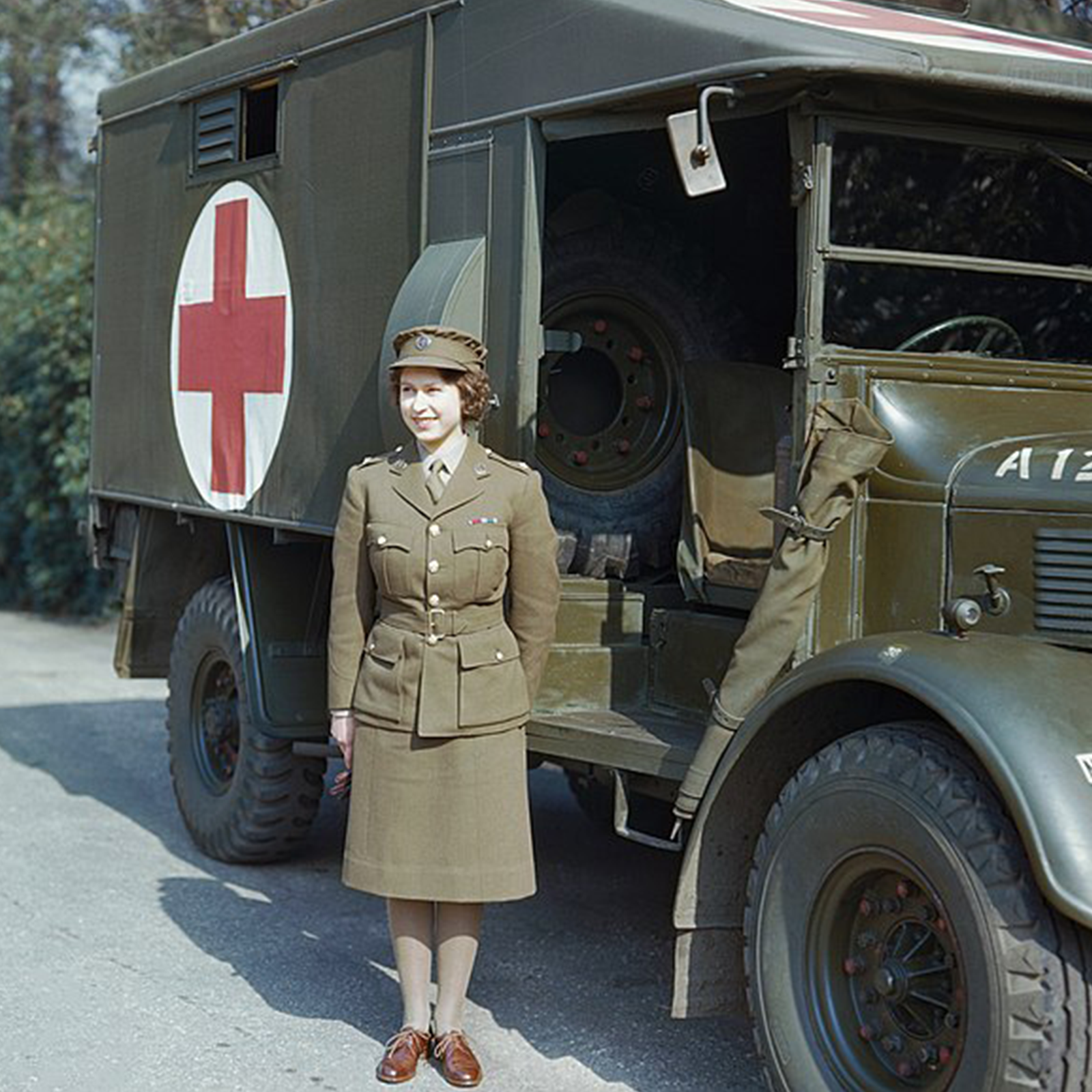 Zdjęcie przedstawia królową Elżbietę II w młodym wieku. Widoczna jest dziewczyna ubrana w zielony mundur składający się z marynarki przepasanej paskiem, spódnicy do kolana, czapki z daszkiem. Dziewczyna stoi przed wojskowym ambulansem, patrzy w prawo i uśmiecha się.