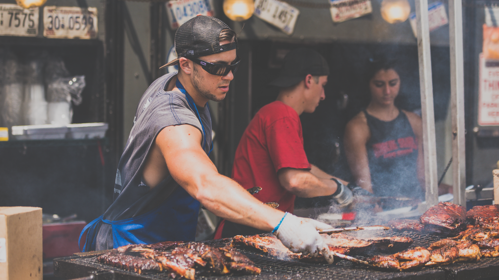Zdjęcie przedstawia na pierwszym planie mężczyznę w ciemnych okularach i koszulce bez rękawów. Na głowie ma czapkę założoną daszkiem do tyłu. Mężczyzna stoi przy ogromnym grillu i przewraca szczypcami mięso. Na drugim planie widoczne są dwie osoby.