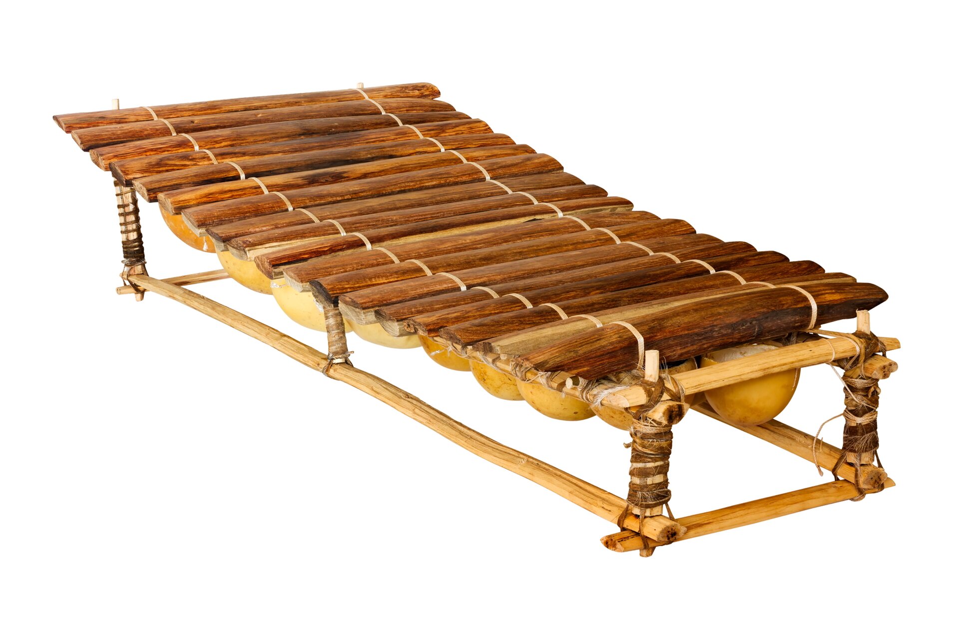 Fotografia przedstawia instrument muzyczny o drewnianej konstrukcji. Podstawa przypomina ławkę o cienkiej desce zamiast siedzenia, następnie z lewej strony obecne są dwa pionowe wystające elementy owinięte materiałem. Pomiędzy nimi umieszczono podłużnie brązowy przedmiot o podłużnym, owalnym kształcie. Następnie, na lewo od niego jest jeszcze osiem podobnych, coraz mniejszych przedmiotów. Ułożono na nich połączone rzemykiem cienkie deseczki, tworzące delikatny łuk.