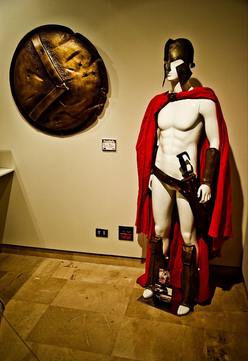 Zdjęcie przedstawia wyposażenie żołnierza spartańskiego z filmy pod tytułem 300. Na męskim manekinie znajdują się: spiżowy hełm (zakrywający głowę, uszy i nos), czerwona peleryna, stalowe ochrony przedramienia, skórzany pas z pochwą i umieszczonym w niej mieczem, stalowe nagolennice. Nieopodal manekina, na ścianie, zawieszona jest duża, okrągła, spiżowa tarcza obronna z literą lambda. Na podstawie informacji zdobytych w lekcji omów, w jaki sposób byli wyposażeni żołnierze spartańscy walczący pod Termopilami.