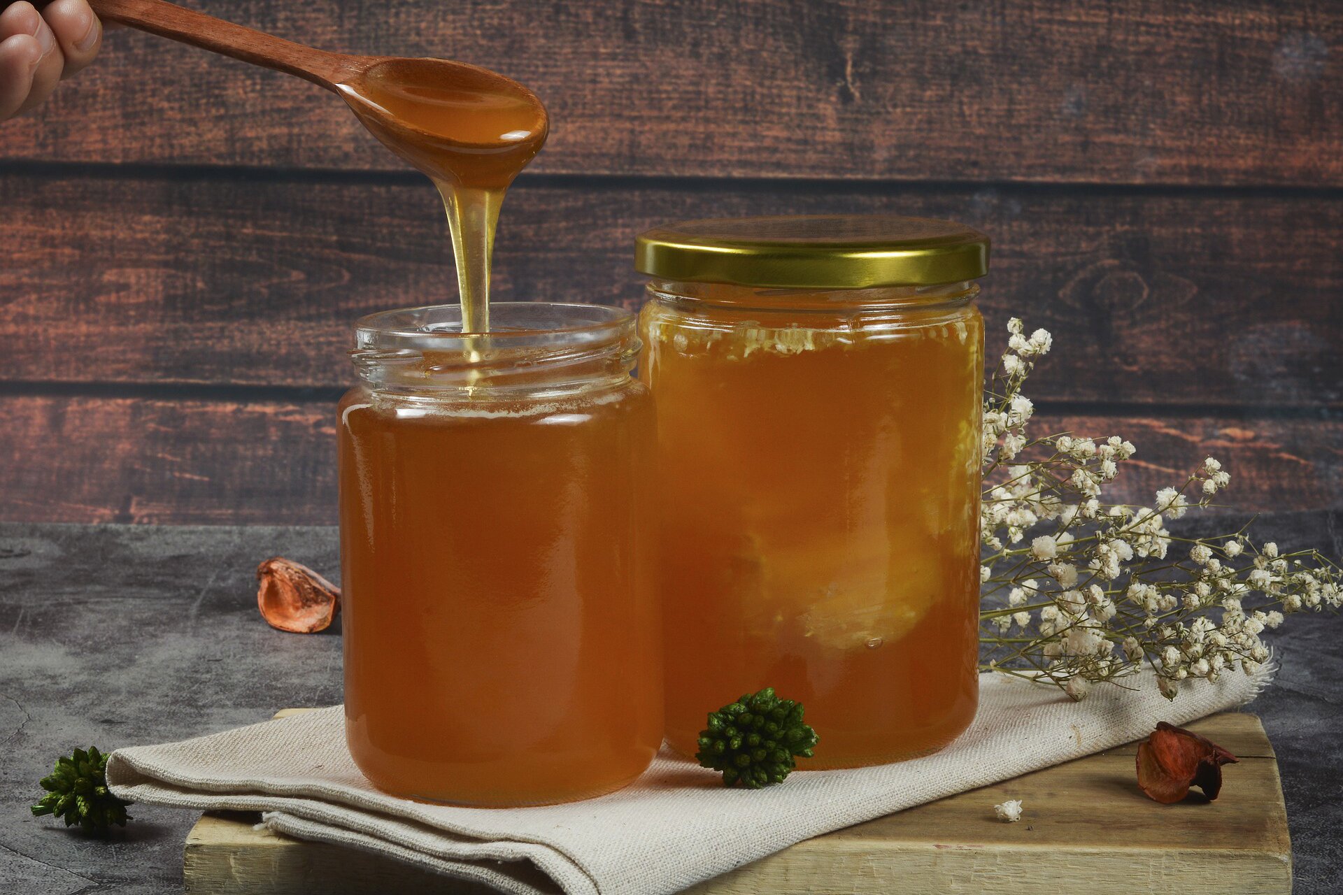 Na planszy widoczne są dwa szklane, przeźroczyste słoje, jeden z żółtą nakrętką, na beżowym materiale, w których znajduje się ciemno‑żółta, gęsta ciecz. Nad jednym słojem widoczna jest drewniana łyżka, oblepiona miodem. Wokół nich widoczne są czarne, białe i czerwone kwiaty.