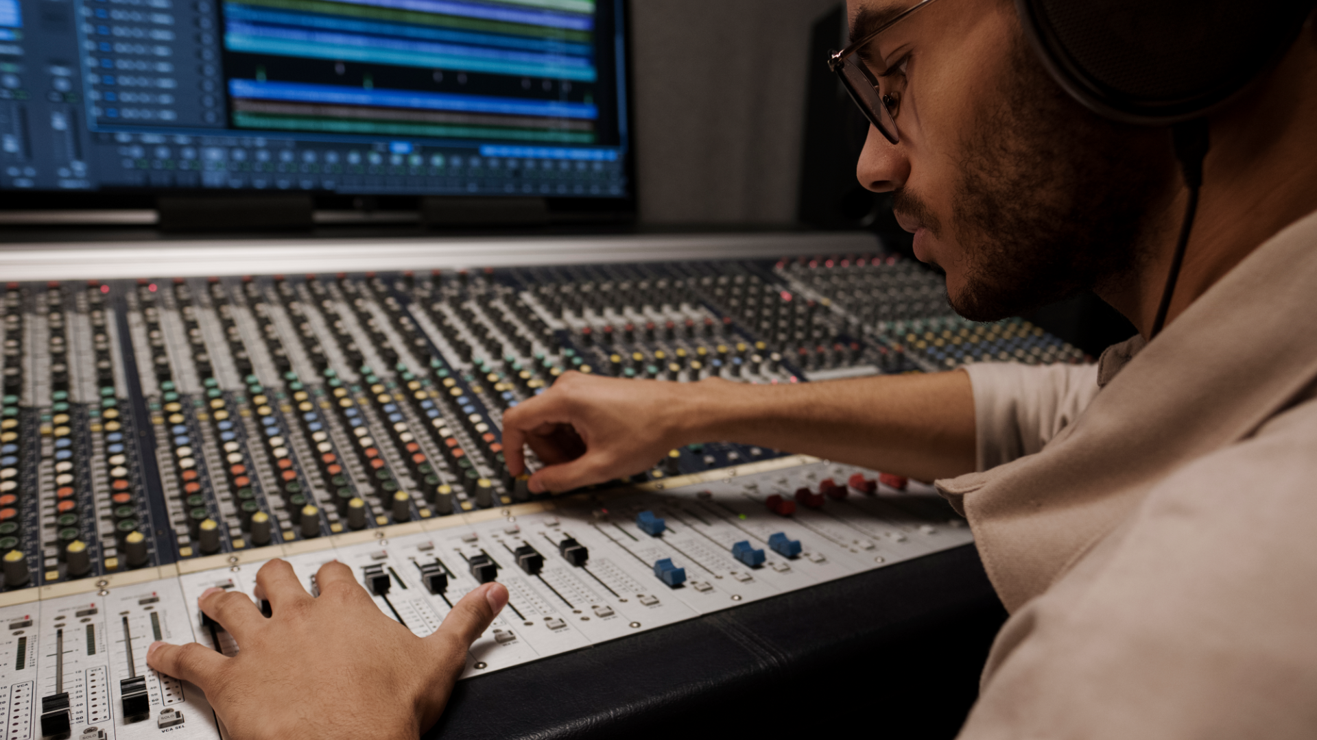 Zdjęcie przedstawia mężczyznę, operatora dźwięku. Mężczyzna jest pochylony nad konsolą pełną przycisków i pokręteł. Przed nim znajduje się monitor. Mężczyzna na uszach ma słuchawki.