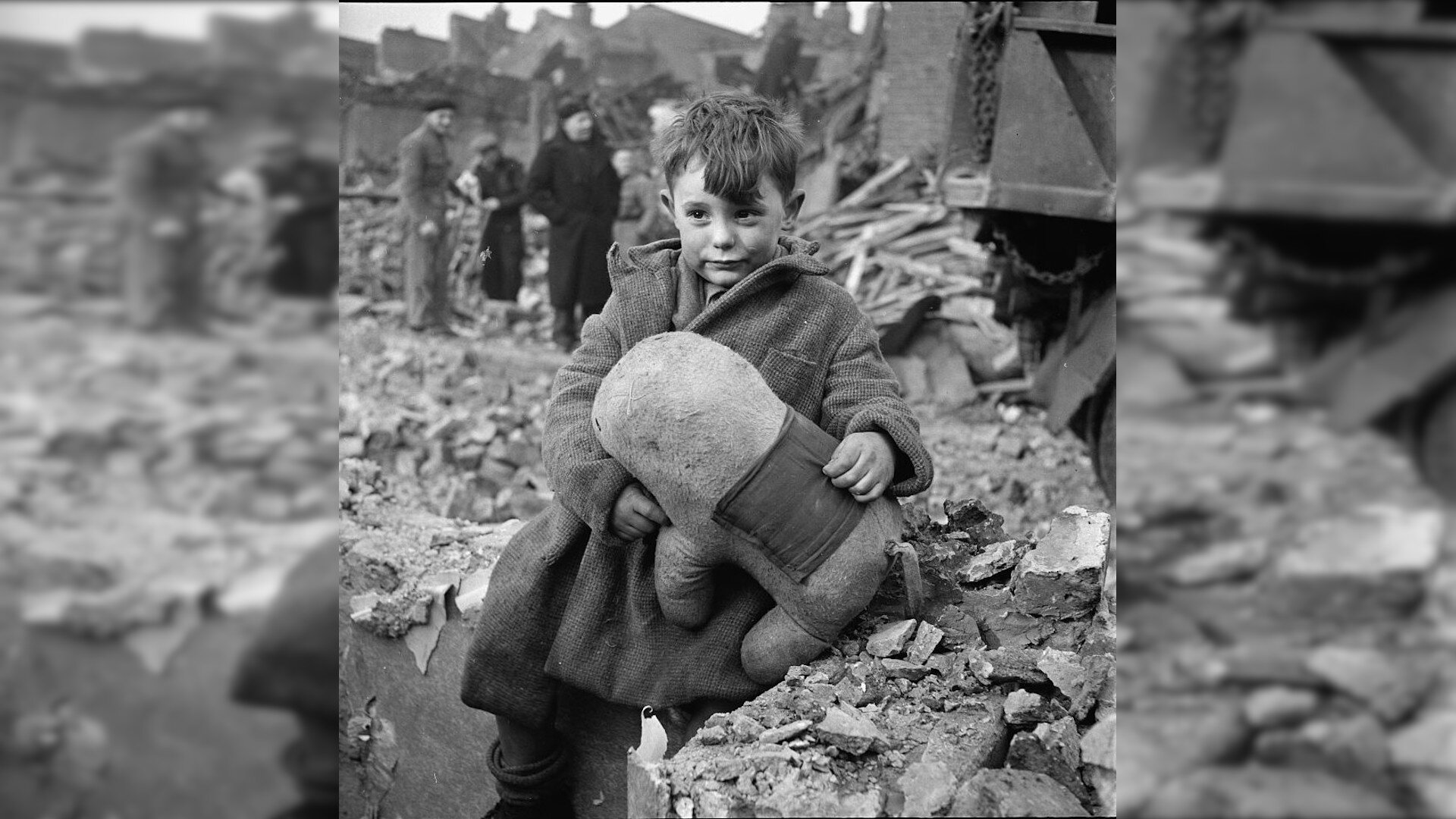 Zdjęcie przedstawia małego chłopca w grubym płaszczu, który trzyma w rękach dużą, pluszową zabawkę. W tle znajdują się inni ludzie oraz gruz i kamienie.