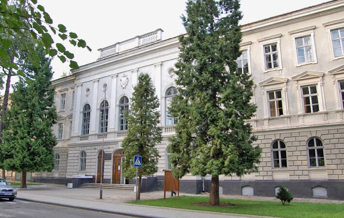 Kliknij, aby powiększyć Kolorowa fotografia przedstawia długi dwupiętrowy budynek o płaskim dachu. Przed nim wąski chodnik i trawnik. Na trawniku rośnie okazała tuja, po jej lewej stronie – dwie kolejne. Pomiędzy nimi znajduje się wejście do budynku, prowadzą do niego trzy szerokie schody. Drzwi są łukowate, drewniane w naturalnym kolorze. Na lewo od nich kolejne, nieco węższe. Elewacja jest utrzymana w jasnych kolorach. Na poziomie parteru biniowanie wokół półokrągłych okien. Okna na pierwszej kondygnacji wykończone pilastrami i w górnej części tympanonami. Na dachu nad głównym wejściem kamienna balustrada.