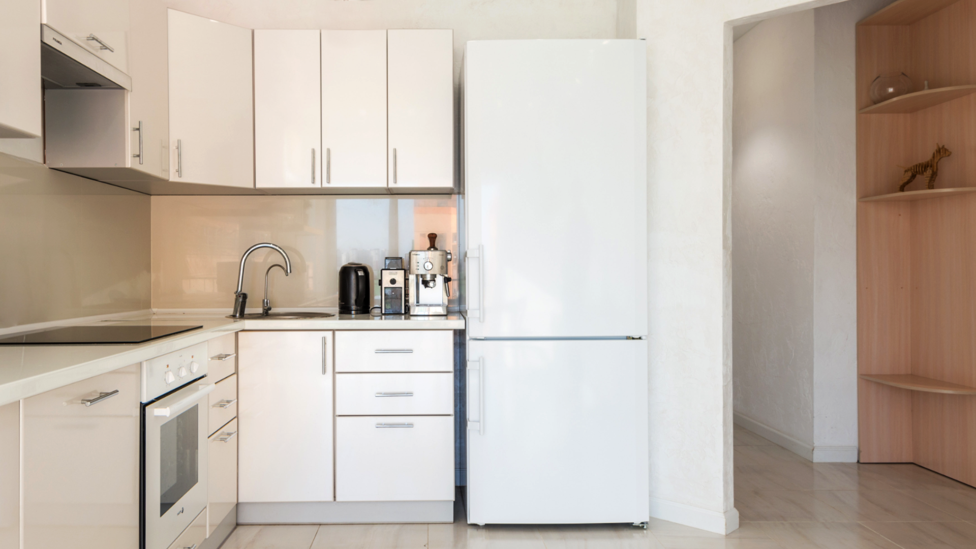 Zdjęcie przedstawia aneks kuchenny. Po prawej stronie stoi lodówka. Na lewo od lodówki znajduje się zlew. Na blacie stoi czajnik oraz ekspres do kawy. Na lewo od zlewu znajduje się piekarnik i płyta grzewcza.