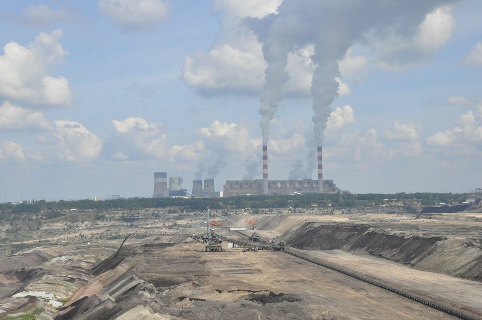 Zdjęcie przedstawia kopalnię odkrywkową węgla brunatnego oraz elektrownię w Bełchatowie. Na pierwszym planie widoczny jest płaski, szeroki teren, na którym pracują maszyny budowlane. W tle znajduje się fabryka z kilkoma niższymi, szerokimi oraz dwoma wysokimi, biało‑czerwonymi kominami, z których w niebo lecą kłęby dymu.